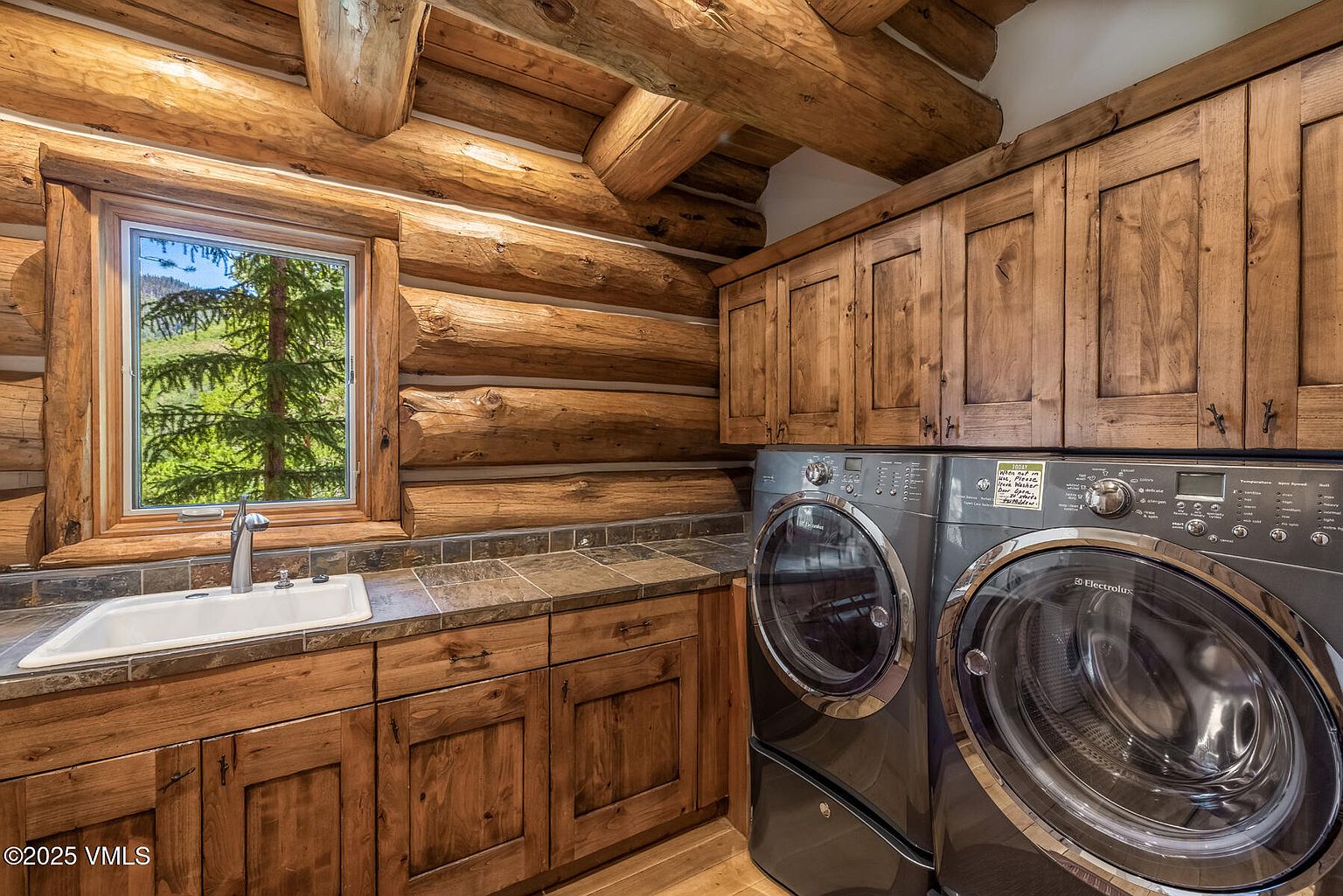 This laundry room features a rustic log cabin design with exposed wooden beams and walls. Modern gray front-loading washer and dryer units are installed beneath wooden cabinets and a tiled countertop with a sink. A window provides natural light and a view of the outdoors, blending rustic charm with modern convenience.