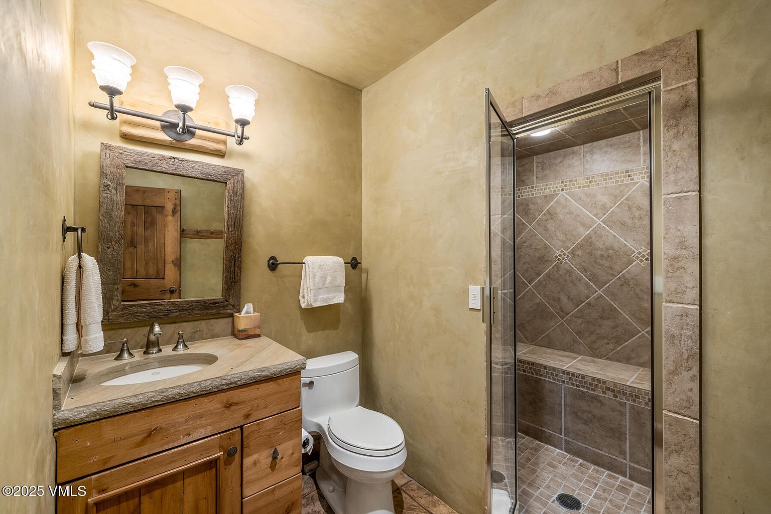 This is a bathroom featuring a rustic vanity with a stone countertop and a wooden-framed mirror. The walls have a textured, earthy tone, and there's a toilet next to the vanity. A glass-enclosed shower with tiled walls and a built-in bench is visible on the right, creating a cozy and inviting space.