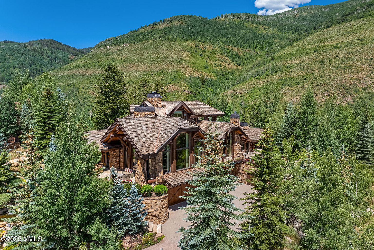 This aerial view showcases a luxurious, multi-story home nestled among lush greenery and mountains. The house features a stone exterior, a wood shingle roof, and multiple chimneys, blending seamlessly with its natural surroundings. A driveway leads to the garage, and mature trees provide privacy and shade, creating an idyllic and secluded estate.