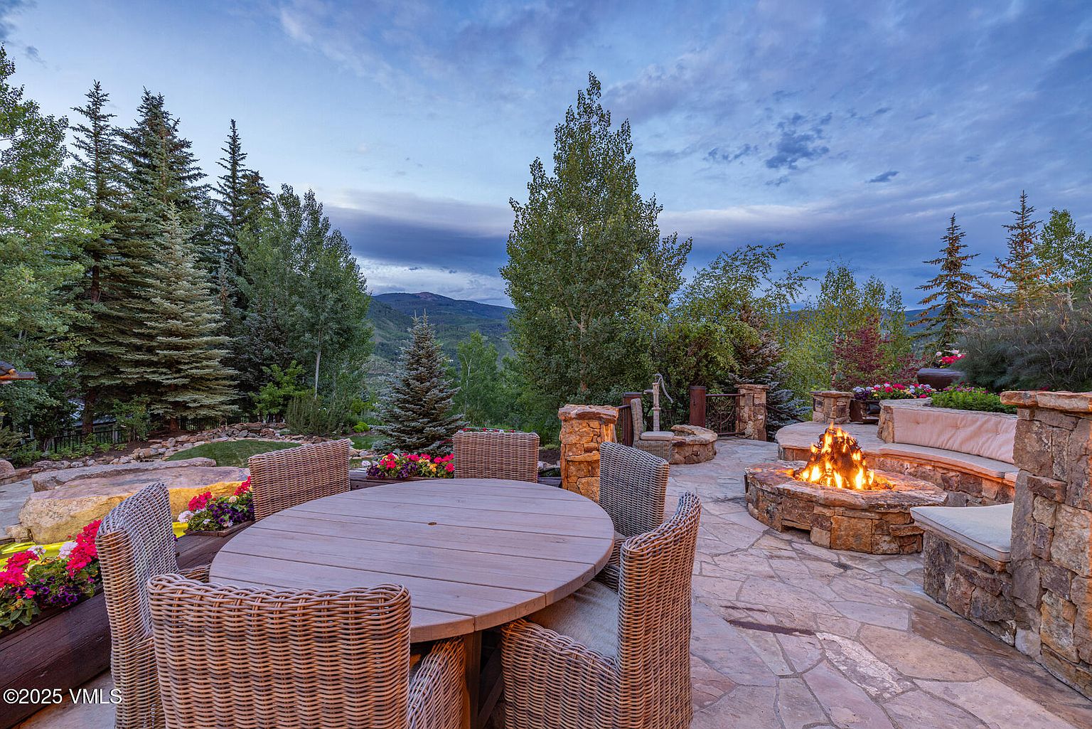 This image showcases a luxurious outdoor patio area, featuring a stone fire pit with flames, a round wooden dining table surrounded by wicker chairs, and a built-in stone bench with cushions. The patio is paved with flagstone and is surrounded by lush greenery, including trees and flowering plants, creating a serene and inviting atmosphere. The perspective is a wide shot, capturing the entire patio area and the surrounding landscape, emphasizing the spaciousness and natural beauty of the property.