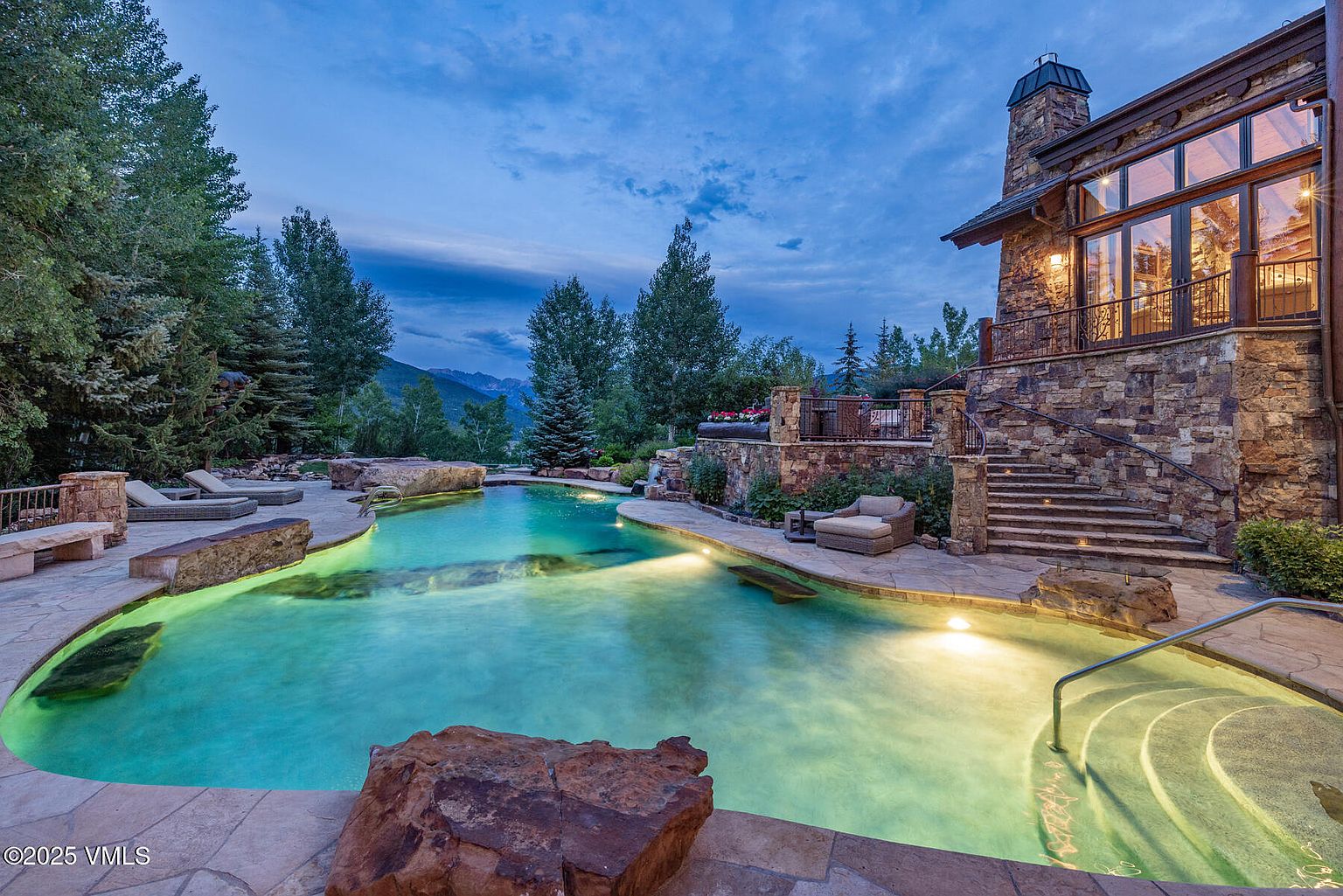 This image showcases a luxurious outdoor pool and spa area at dusk, featuring a uniquely shaped pool with built-in rock features and underwater lighting. The pool is surrounded by stone paving and lush landscaping, with a stone-clad house visible in the background, complete with a balcony and large windows. The scene exudes a sense of tranquility and high-end living.