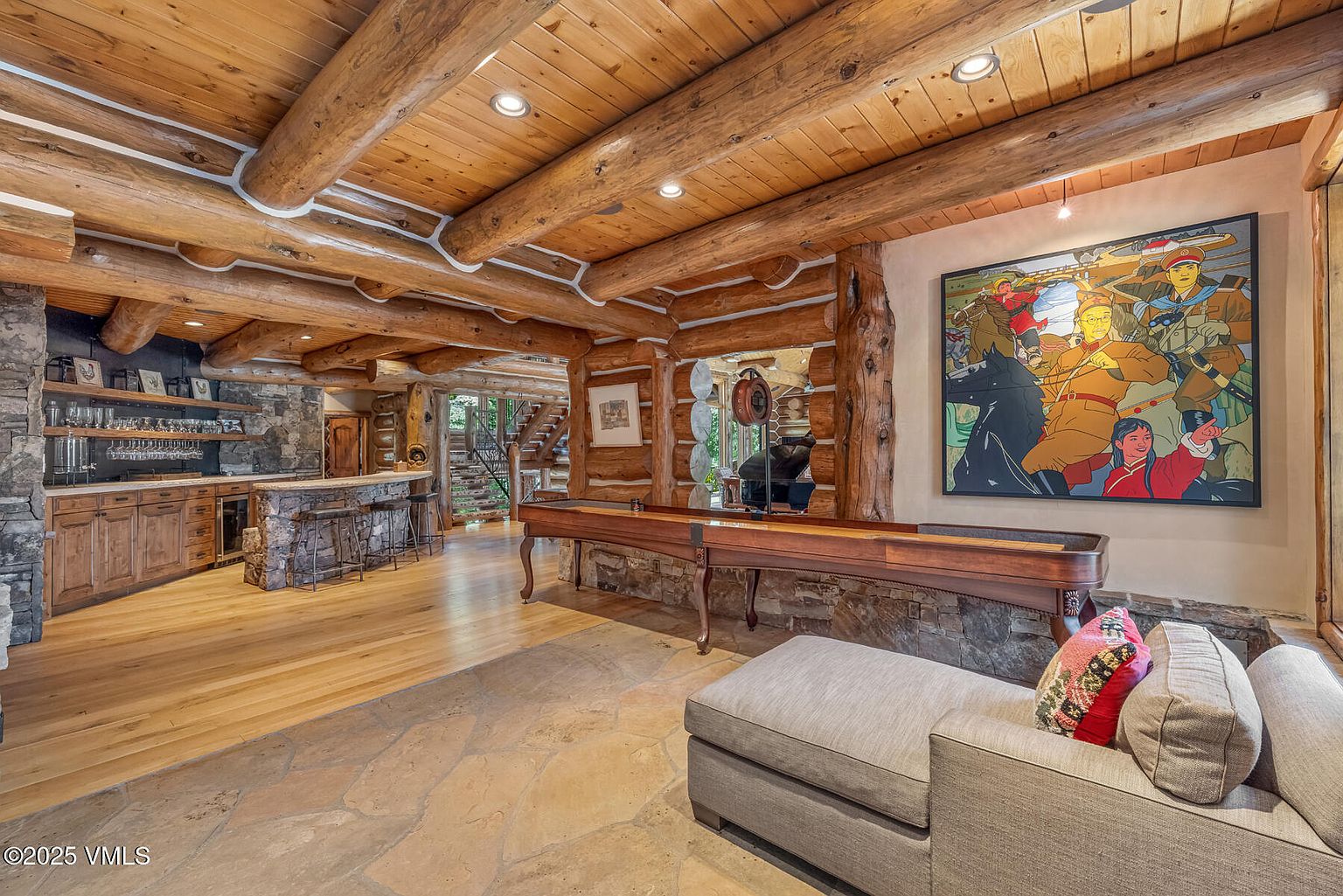 This interior shot showcases a rustic living room with log beam ceilings and stone accents. A shuffleboard table sits prominently in the foreground, leading to a bar area with wooden cabinets and open shelving. The room is furnished with a comfortable sofa and features a large, colorful painting on the wall, creating a warm and inviting atmosphere.