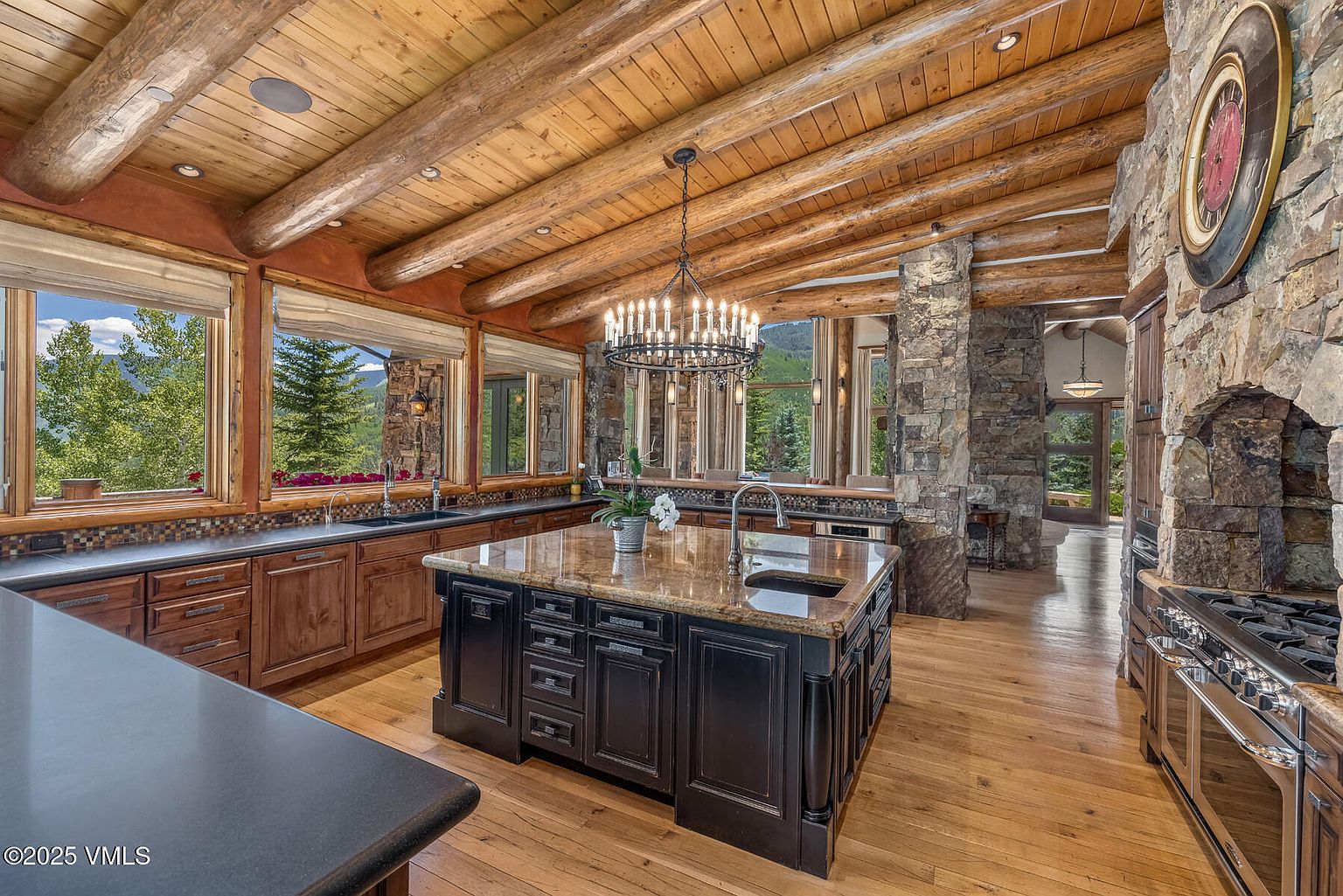 This is a spacious kitchen featuring a rustic design with wooden beams on the ceiling and stone accents on the walls. A large kitchen island with a granite countertop and dark cabinetry is centrally located, complemented by wooden cabinets along the walls. Large windows offer views of the surrounding landscape, enhancing the natural and inviting atmosphere of the space.