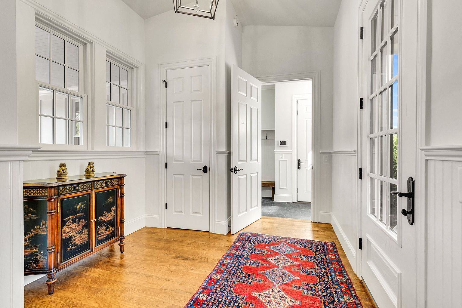 This bright and elegant entryway hallway features crisp white wainscoting, hardwood floors, and a vibrant, traditional-style area rug. The space is accented by a decorative, dark-wood cabinet with intricate oriental-inspired artwork and is illuminated by a classic lantern-style pendant light. The perspective looks down the corridor, showcasing multiple white paneled doors and a glimpse into a secondary mudroom area, creating a welcoming and sophisticated transition space.