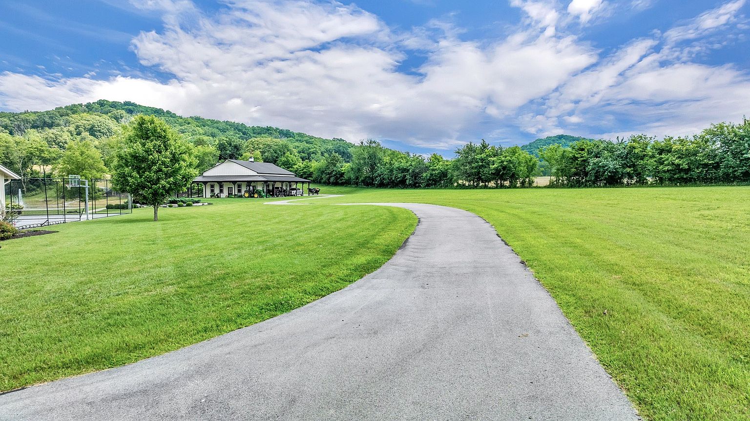 This expansive property features a winding paved driveway leading toward a large, well-maintained barn-style outbuilding set against a backdrop of lush green hills. The scene is characterized by a vast, manicured lawn, mature trees, and a clear view of a private basketball court to the left. The perspective is captured from a ground-level vantage point, emphasizing the open, serene, and spacious nature of the rural landscape.