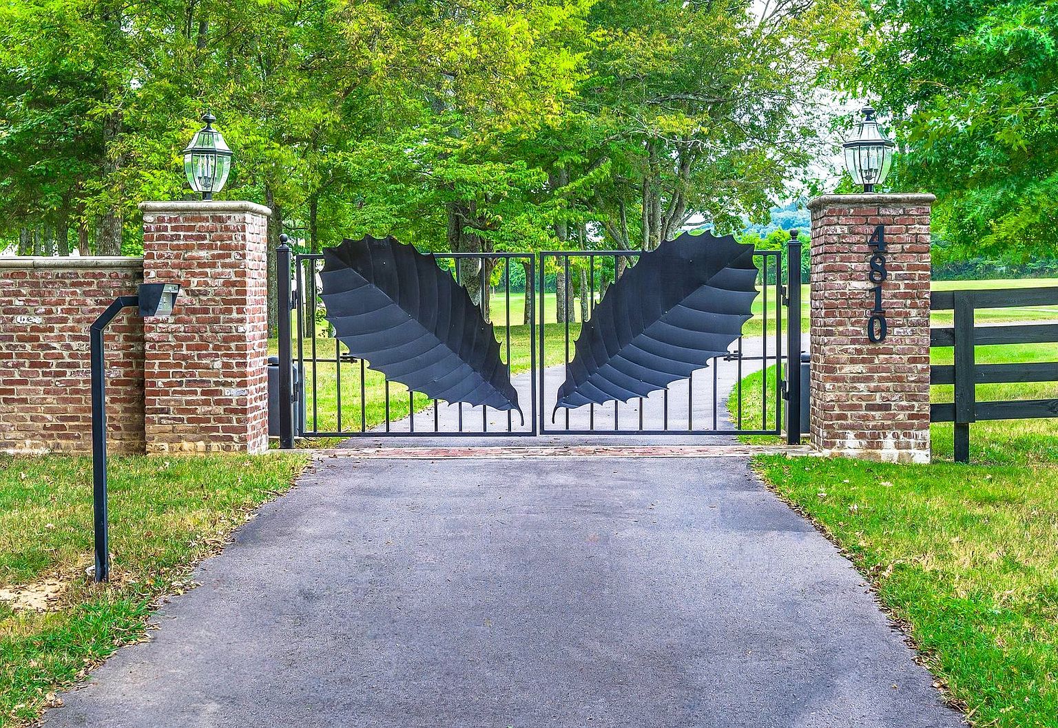 This image showcases an impressive entryway featuring a custom metal gate with leaf-shaped designs, flanked by brick pillars topped with lantern-style lights. A paved driveway leads through the gate, framed by well-maintained lawns and mature trees in the background. The overall impression is one of elegance and privacy.