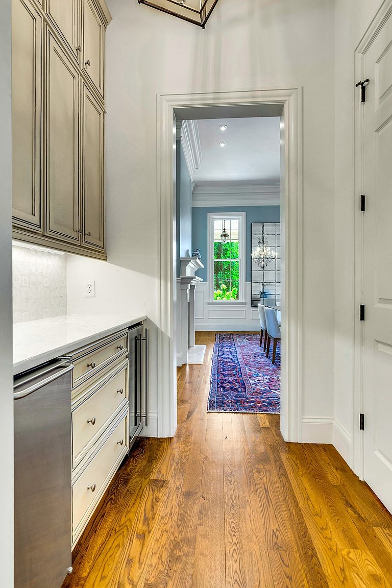 The image showcases a hallway with hardwood flooring leading into a dining room. On the left, there are built-in cabinets and drawers with a light countertop, while on the right, a white door with black hardware is visible. The dining room features a blue accent wall, a window with natural light, and a chandelier, creating an inviting and elegant atmosphere.