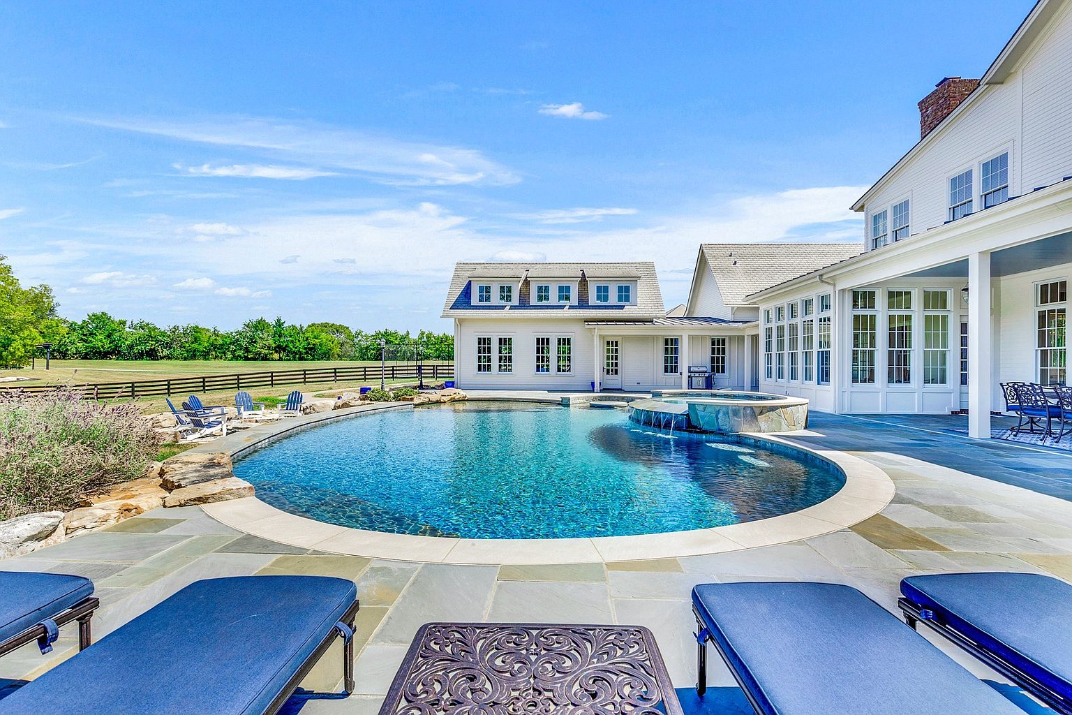This stunning backyard oasis features a large, custom-shaped swimming pool with an integrated stone-clad spa that spills water into the main pool. The expansive stone patio is furnished with comfortable lounge chairs, offering a perfect vantage point for enjoying the serene, open landscape and the elegant white farmhouse-style architecture of the home. The scene captures a bright, sunny day, emphasizing a luxurious and relaxing outdoor living environment.