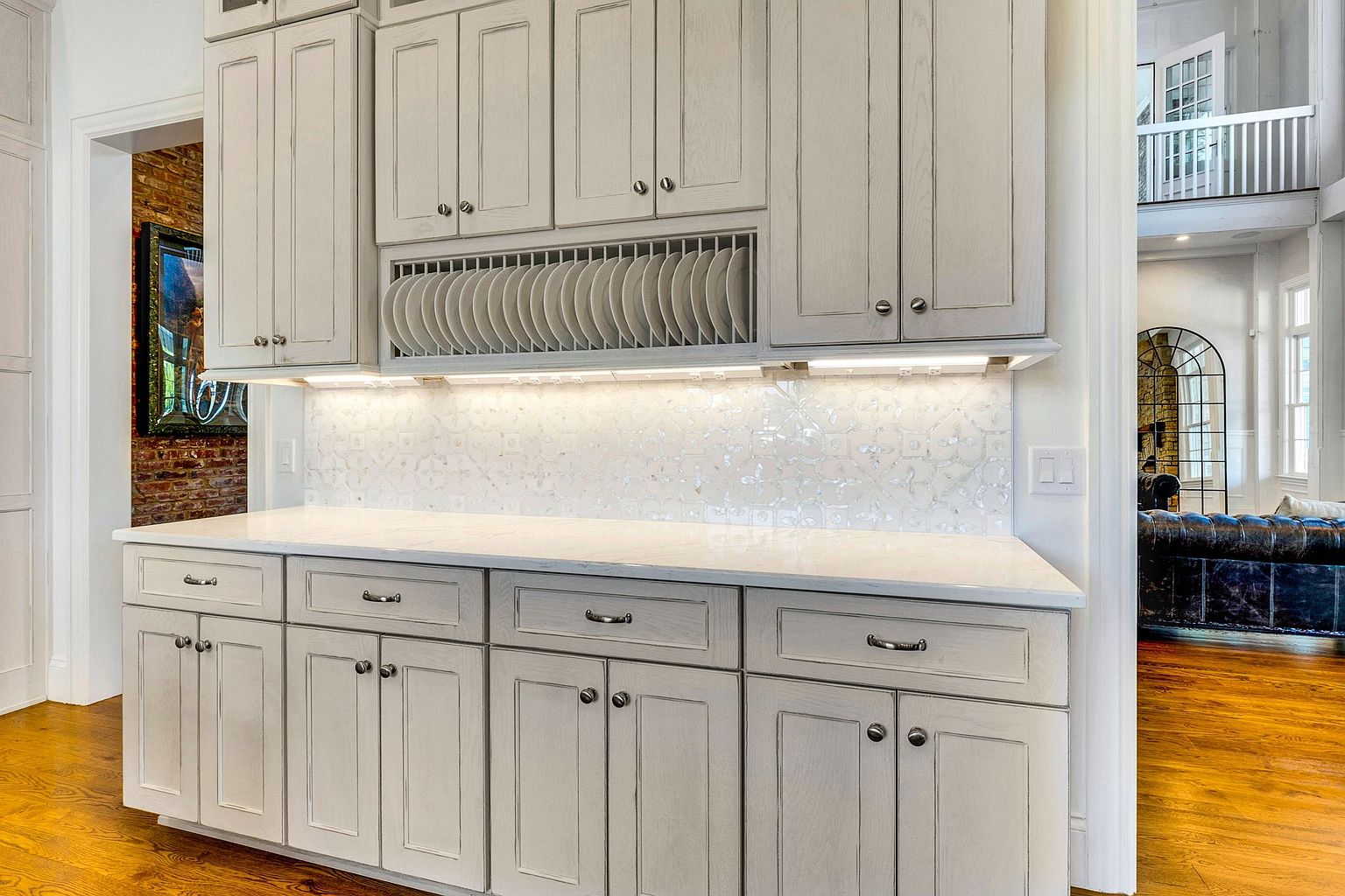 This elegant kitchen area features light-toned, distressed-finish cabinetry with a built-in plate rack and under-cabinet lighting. The space is anchored by a pristine white countertop and a decorative, glossy patterned backsplash, creating a sophisticated and bright atmosphere. The perspective captures the cabinetry and counter space, with a glimpse into an adjacent living area featuring a brick wall and a leather sofa, suggesting an open-concept layout.