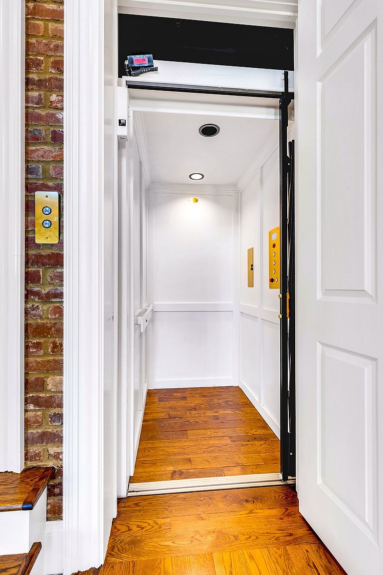 This image captures an open residential elevator framed by a classic brick wall and white trim, creating a sophisticated transition between floors. The interior of the elevator features clean white paneling, warm hardwood flooring, and recessed lighting, offering a seamless blend of traditional and modern design. The perspective is eye-level, looking directly into the elevator car from the adjacent hallway.