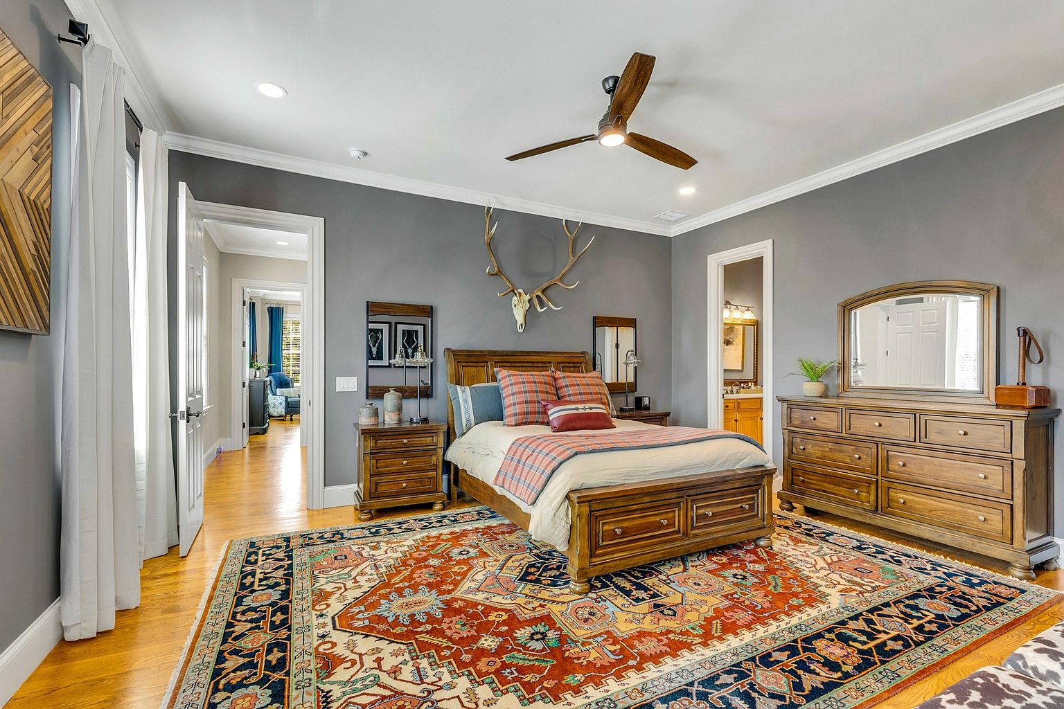 This spacious primary bedroom features a warm, rustic aesthetic with a wooden bed frame, matching dresser, and a large, colorful patterned area rug. The room is anchored by a prominent deer antler wall mount above the bed, set against a sophisticated grey wall color that contrasts with the hardwood floors. The perspective is a wide-angle shot from the foot of the bed, showcasing the room's layout and its connection to the adjacent hallway and bathroom.