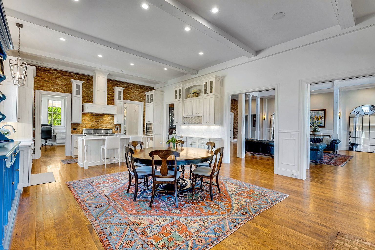 This is a spacious, open-concept kitchen and dining area featuring white cabinetry, a brick accent wall, and hardwood floors. A large round dining table sits on an ornate rug, creating a focal point. The kitchen island provides additional seating, and the space flows seamlessly into adjacent living areas, offering a sense of connectivity and modern living.