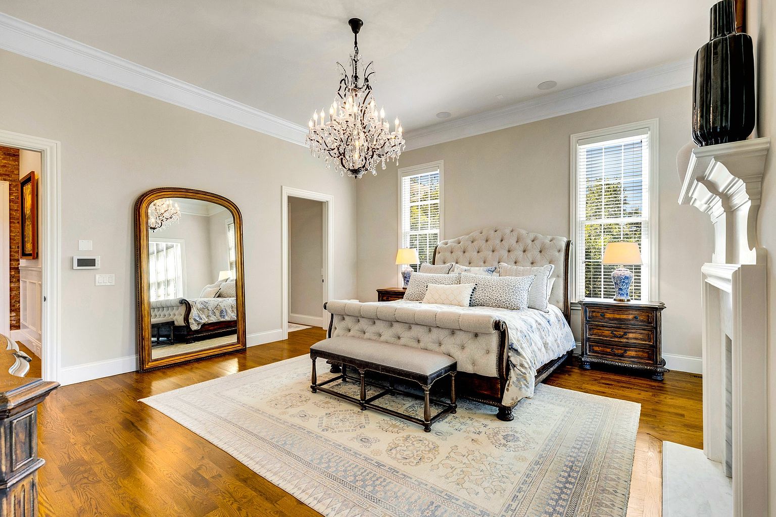 This is a luxurious primary bedroom featuring a tufted headboard bed with a bench at the foot, complemented by a detailed area rug. The room is illuminated by a chandelier and natural light from the windows, with bedside tables and lamps adding to the ambiance. A large mirror leans against the wall, reflecting the room's elegance.