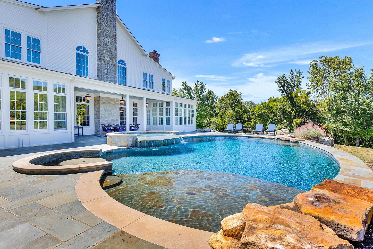 This stunning backyard oasis features a custom-designed swimming pool with an integrated raised spa that spills over into the main pool area. The scene is framed by a large stone patio, natural rock accents, and a beautiful white multi-story home with expansive windows. The bright, sunny atmosphere and lush green trees in the background create a luxurious and inviting resort-style living space.
