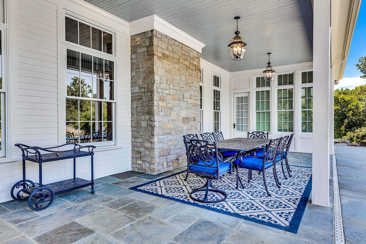 This elegant covered patio features natural stone flooring and a prominent stone fireplace chimney, creating a sophisticated outdoor living space. The area is furnished with a dark metal dining set with vibrant blue cushions, complemented by a matching serving cart and a patterned area rug. The space is framed by white siding, large windows, and classic hanging lanterns, offering a serene view of the surrounding greenery.