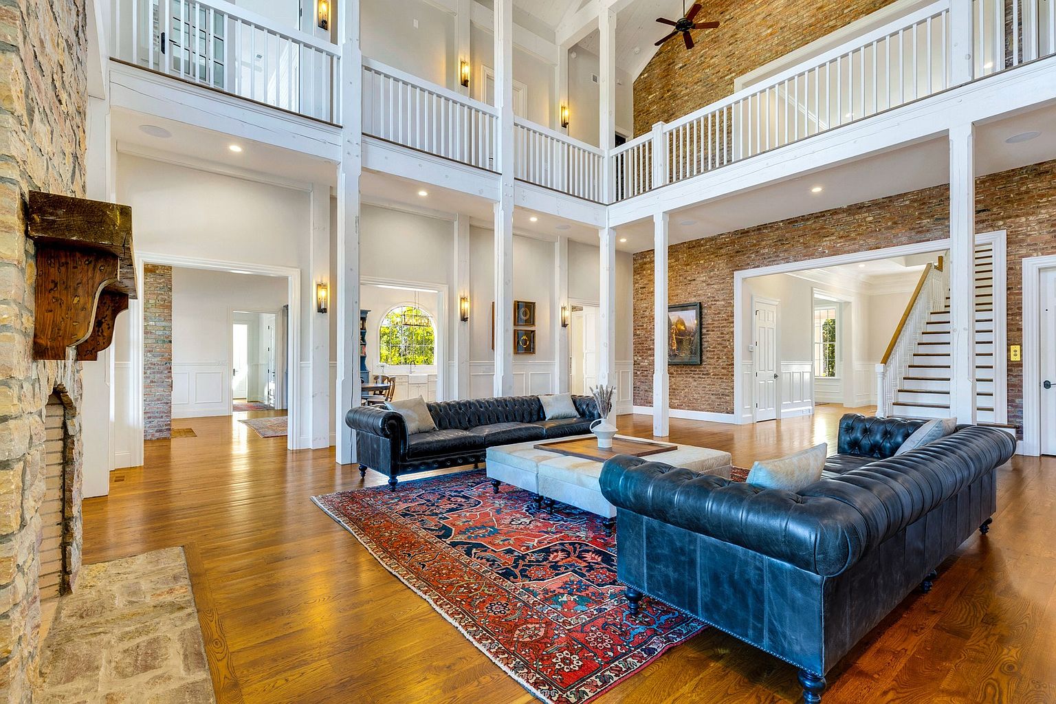 This is a grand living room featuring soaring ceilings, exposed brick accents, and substantial white columns. Two dark leather sofas flank a large ottoman, centered on an ornate rug. The space is bathed in natural light, highlighting the hardwood floors and creating a luxurious, open atmosphere.