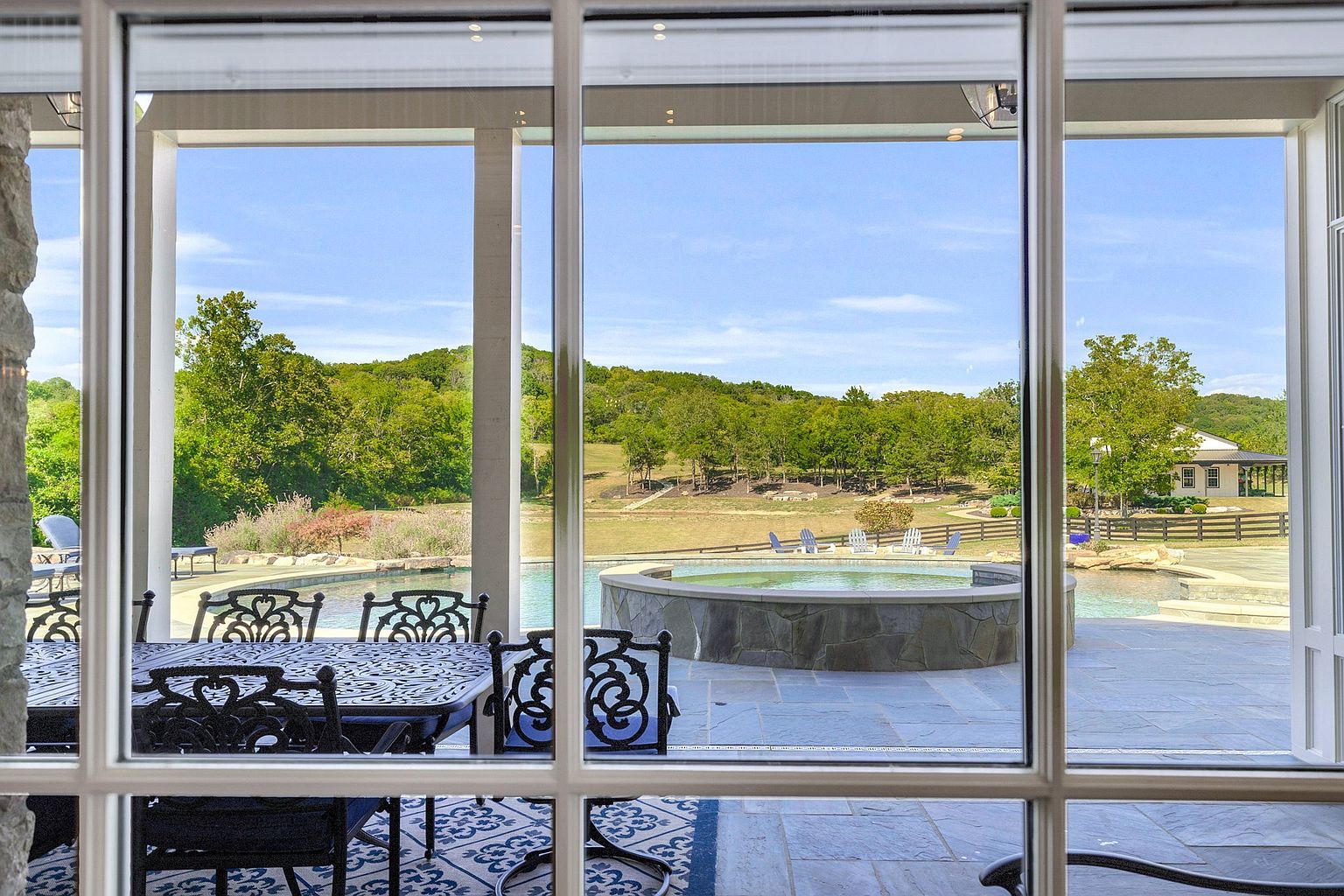 This image showcases a luxurious outdoor pool and spa area, viewed through a large window from an interior space. The pool features a stone-clad spa, surrounded by a spacious patio. Beyond the pool area, a well-manicured lawn extends towards a wooded area, creating a serene and private backyard oasis.