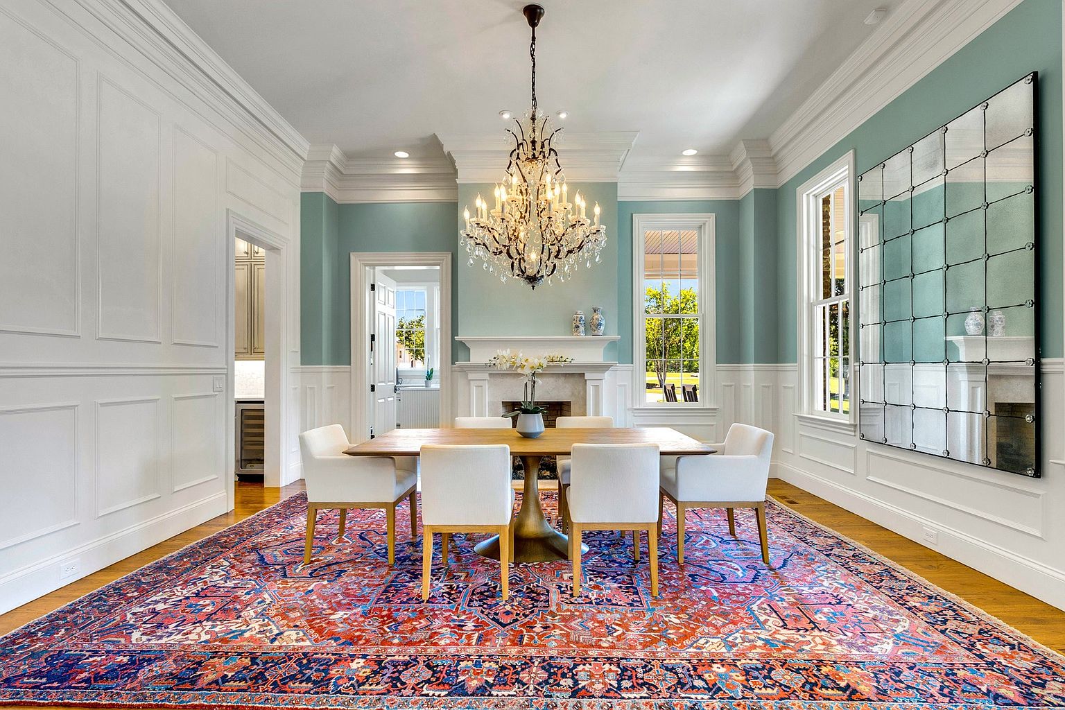 This elegant dining room features a sophisticated blend of traditional and modern design, highlighted by a large, ornate crystal chandelier centered above a wooden dining table with six upholstered chairs. The room is anchored by a vibrant, patterned area rug and features wainscoting, a fireplace, and a large grid-style mirror that enhances the sense of space. The soft blue wall color and natural light streaming through the windows create a bright, inviting, and formal atmosphere perfect for entertaining.