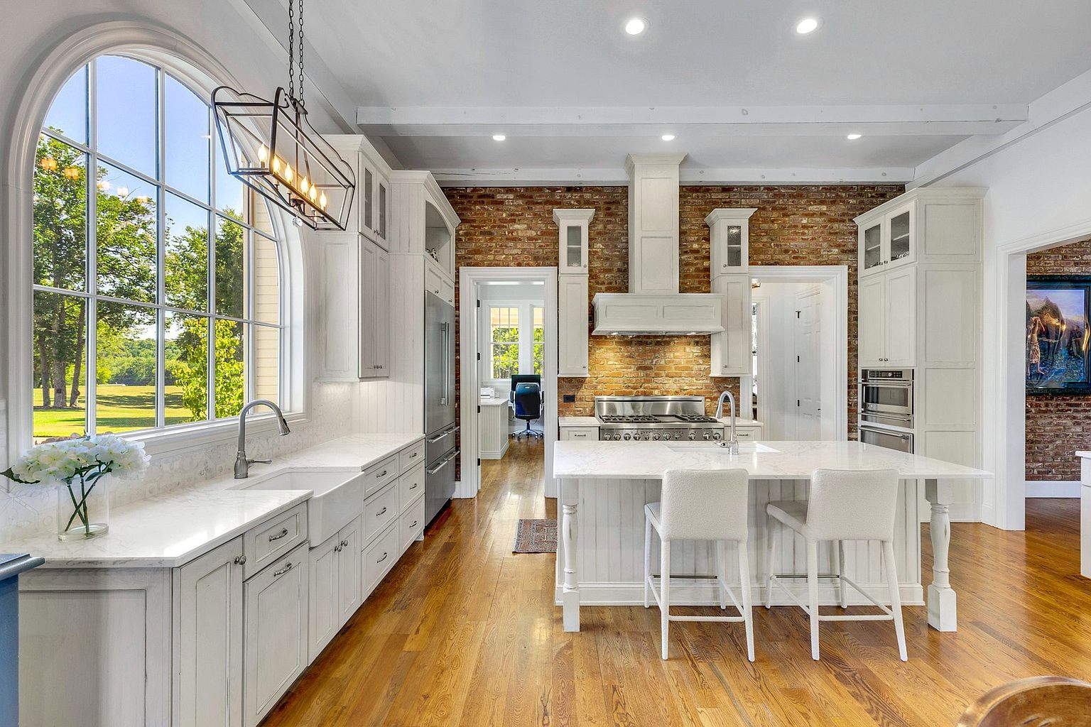 This elegant, high-end kitchen features a blend of rustic exposed brick walls and sophisticated white cabinetry with marble countertops. A large central island with seating serves as a focal point, complemented by a farmhouse sink and a striking arched window that floods the space with natural light. The warm hardwood floors and professional-grade appliances create a luxurious yet inviting atmosphere perfect for culinary enthusiasts.
