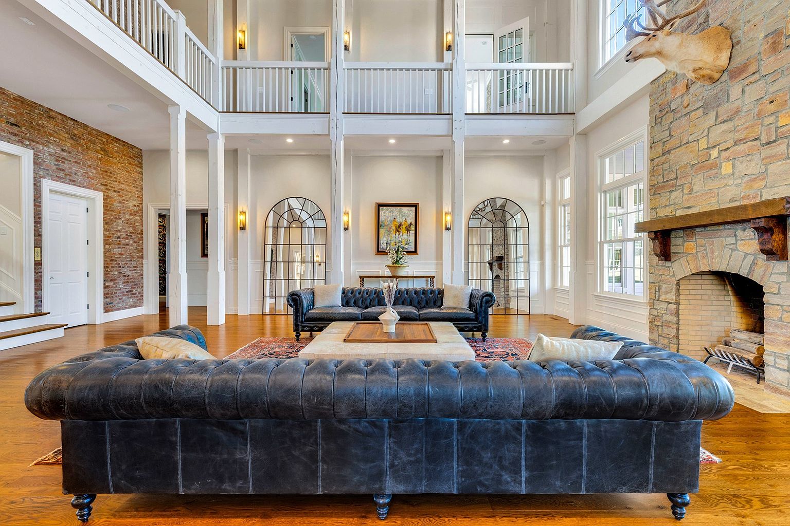 This grand, double-height living room features a sophisticated blend of rustic and refined elements, anchored by a massive stone fireplace adorned with a mounted elk head. The space is defined by two luxurious, dark leather tufted sofas facing each other, separated by a large, light-toned coffee table on a patterned area rug. Architectural details include exposed brick walls, elegant arched mirrors, and a mezzanine balcony with white railings, creating an airy and opulent atmosphere.