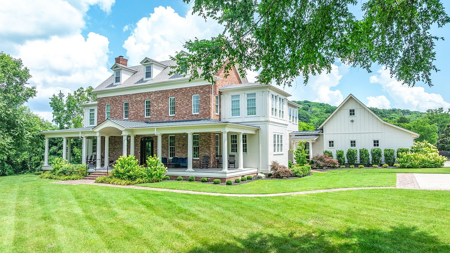 This elegant, multi-story estate features a classic blend of red brick and white siding, accented by a sprawling wrap-around porch supported by white columns. The property is set against a backdrop of lush green trees and a manicured lawn, with a secondary white farmhouse-style structure attached to the side. The bright, sunny day highlights the architectural charm and expansive outdoor living space, creating an inviting and prestigious atmosphere.