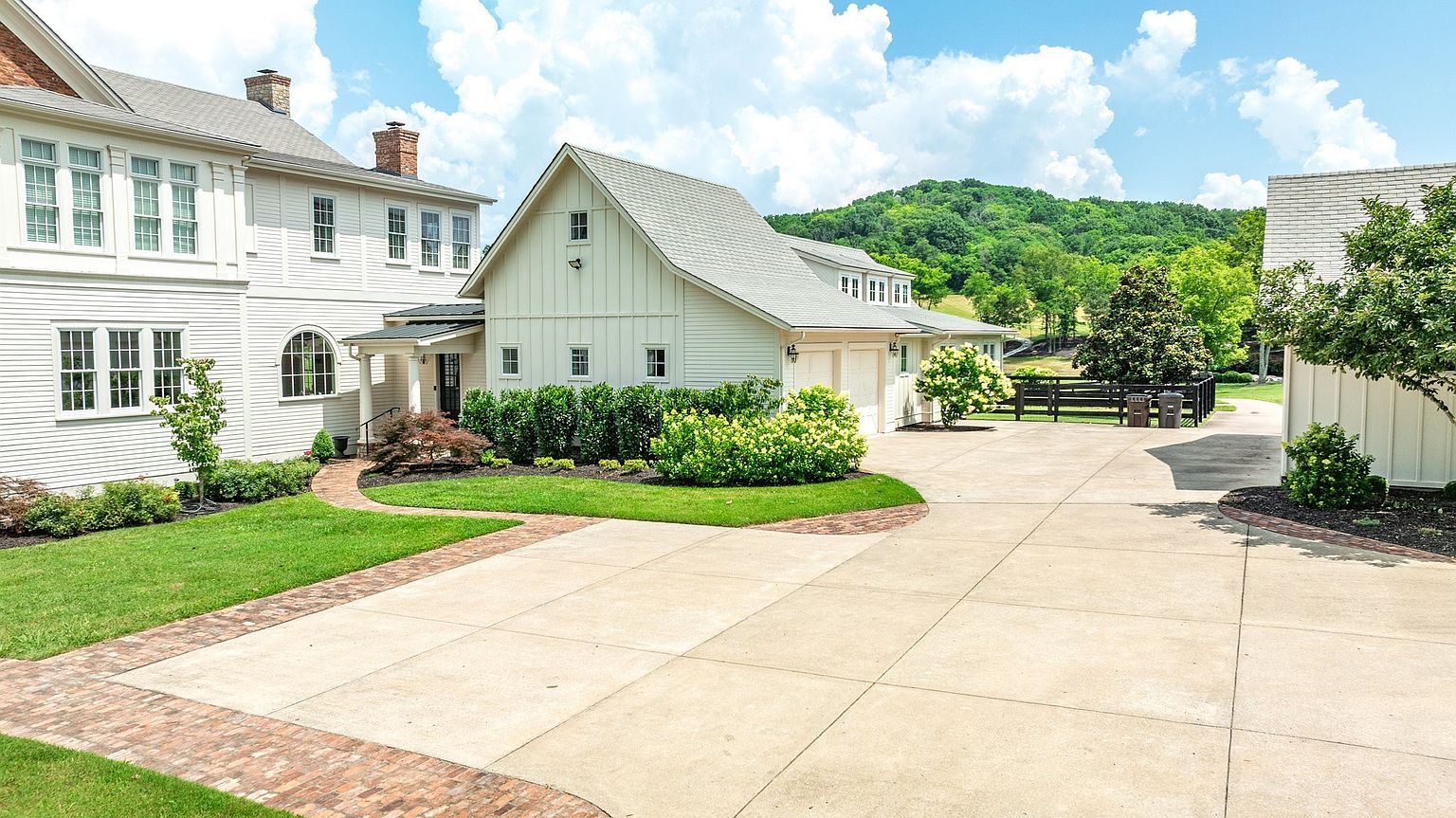 This exterior view showcases a charming, light-colored farmhouse-style home with a spacious concrete driveway and a well-manicured lawn. The property features a multi-car garage, elegant brick-bordered walkways, and lush landscaping, all set against a backdrop of rolling green hills. The perspective captures the inviting transition between the main residence and the garage structure, emphasizing the home's curb appeal and peaceful rural setting.