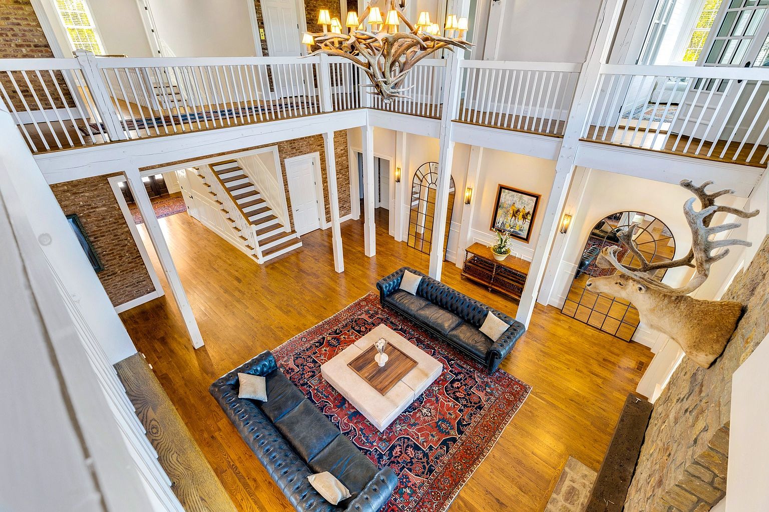 This high-angle, cinematic shot captures a grand, two-story living room characterized by its rustic elegance and open-concept design. The space features a large, ornate antler chandelier, two dark leather tufted sofas arranged around a central square ottoman, and a vibrant, traditional area rug set against warm hardwood floors. A prominent stone fireplace wall adorned with a large taxidermy deer mount anchors the room, while white railings on the mezzanine level provide a clear view down into the inviting, light-filled living area.