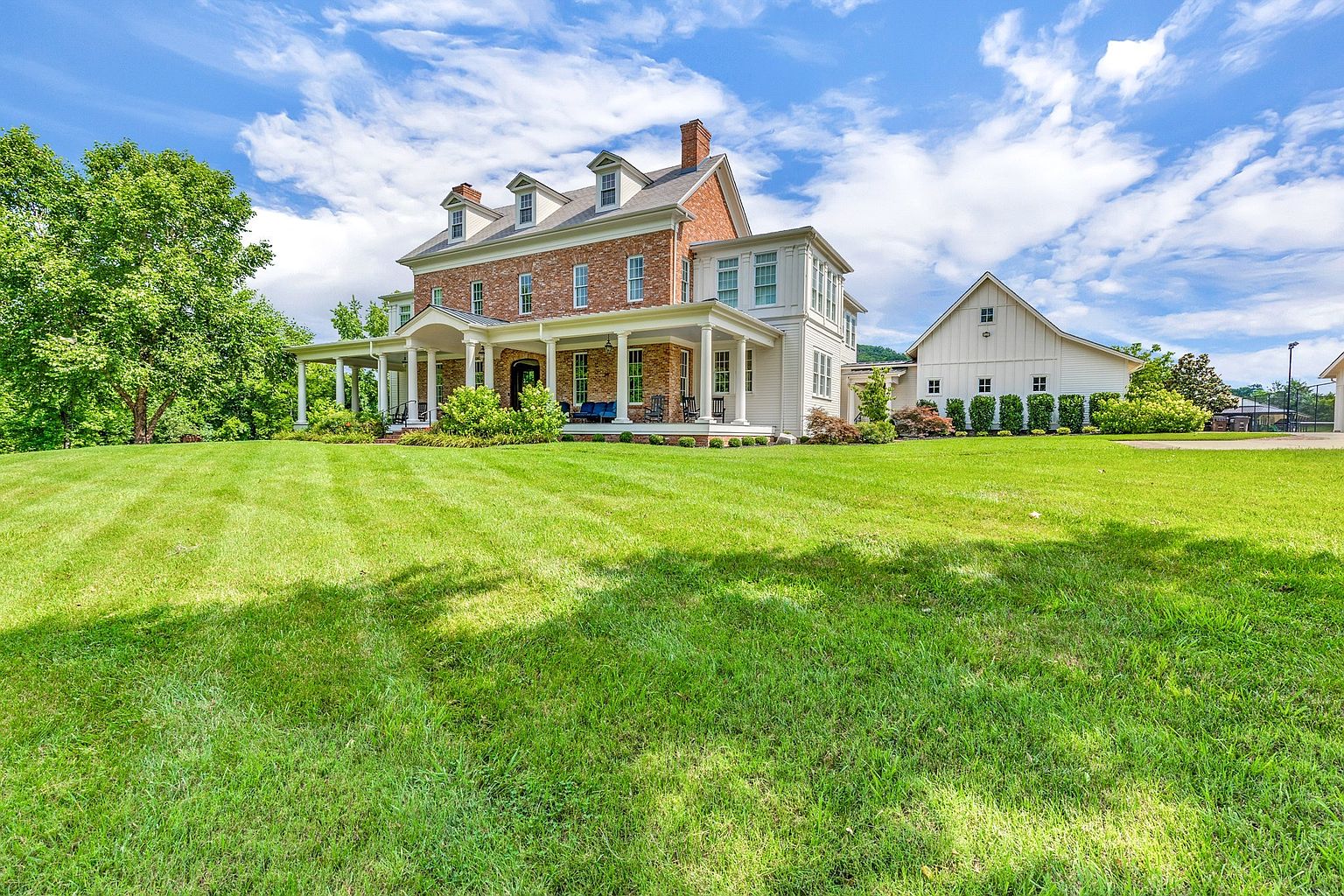 This is a stunning front view of a large, elegant brick house with a sprawling green lawn. The house features a wide, inviting porch with white columns, dormer windows, and a prominent chimney. A white barn-like structure is visible in the background, adding to the property's charm and spaciousness. The perspective is a wide shot, capturing the grandeur of the estate under a bright, partly cloudy sky.