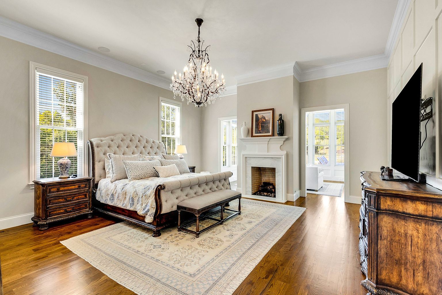 This is a luxurious primary bedroom featuring a tufted headboard bed with decorative pillows, a bench at the foot of the bed, and a large area rug. The room includes a fireplace with a marble surround, a chandelier, and large windows providing natural light. The overall impression is elegant and spacious, with a focus on comfort and style.