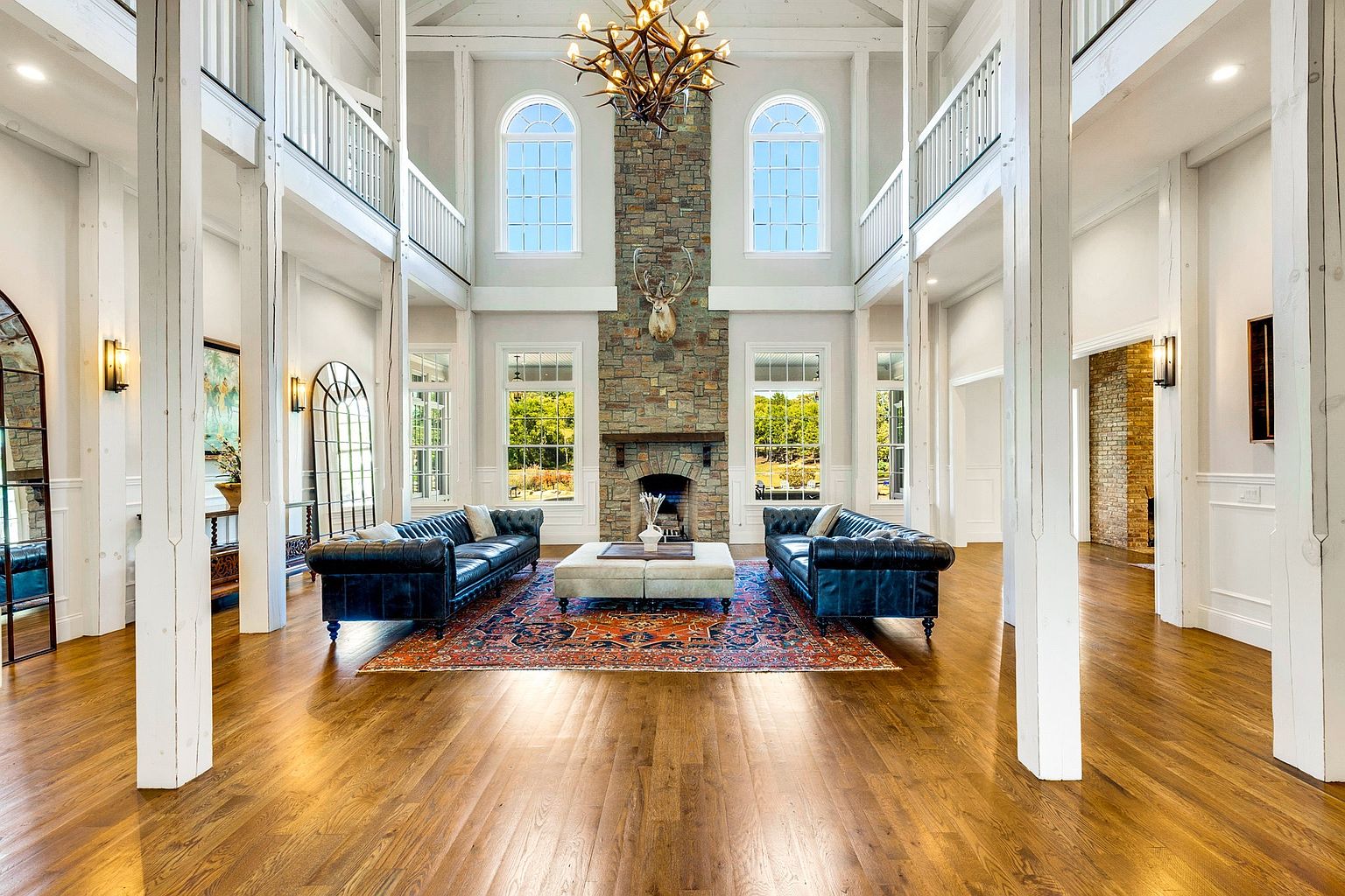 This grand living room features soaring ceilings with exposed beams, supported by large white columns. A stone fireplace with a mounted deer head serves as a focal point, flanked by arched windows and matching black leather sofas. An ornate rug and chandelier add to the room's luxurious and rustic-chic ambiance.