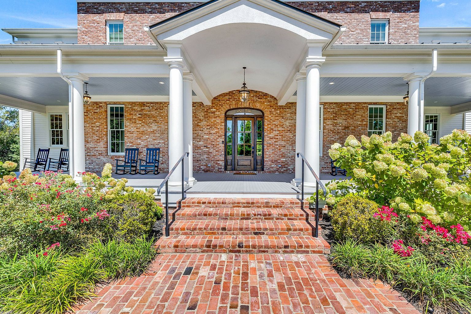 This inviting front entryway features a classic brick facade with a grand covered porch supported by white columns. A symmetrical arrangement of rocking chairs and lush, blooming landscaping creates a warm, welcoming atmosphere, while the central wooden front door serves as a sophisticated focal point. The perspective is a straight-on, eye-level shot that emphasizes the home's traditional charm and curb appeal.