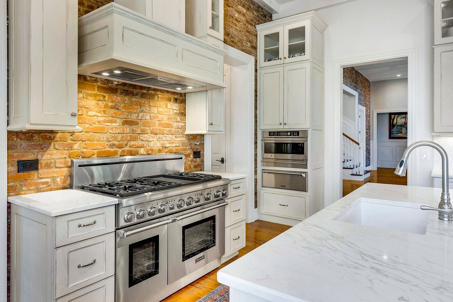 This elegant kitchen features a striking exposed brick wall that contrasts beautifully with the crisp white cabinetry and marble countertops. A professional-grade stainless steel range serves as the focal point, complemented by a built-in wall oven and microwave unit. The perspective captures the functional layout, highlighting the seamless blend of rustic charm and modern luxury.