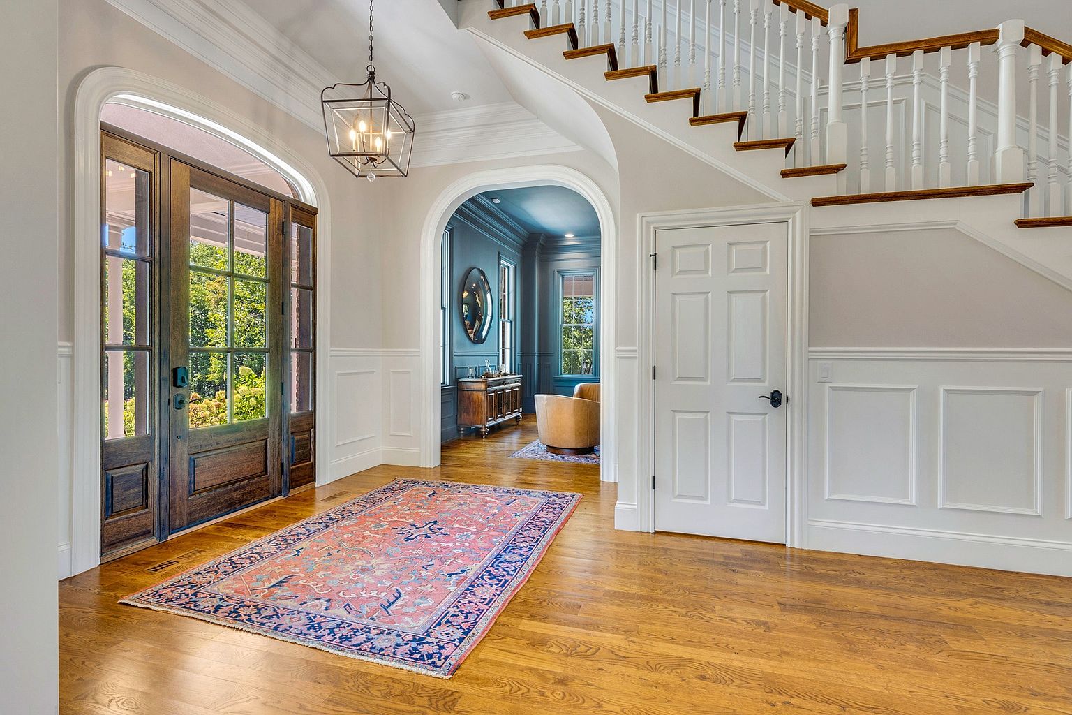 This elegant foyer features a grand staircase with wooden treads and white balusters, set against a backdrop of neutral walls with classic wainscoting. A large, ornate wooden front door with glass panels allows natural light to flood the space, highlighting the rich hardwood flooring and a vibrant, traditional-style area rug. The perspective looks through an arched doorway into a sophisticated, deep-blue room, creating a sense of depth and architectural interest.