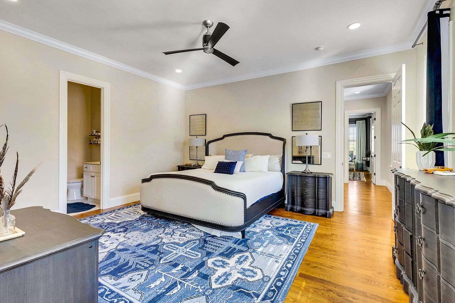 This spacious primary bedroom features a sophisticated upholstered bed frame as the focal point, set against a neutral wall palette and anchored by a vibrant, intricate blue patterned area rug. The room is well-lit with recessed lighting and a modern ceiling fan, while dark wood furniture pieces provide a striking contrast. The perspective captures the bedroom's flow, showcasing an en-suite bathroom entrance on the left and a hallway leading to another room on the right, creating an inviting and airy atmosphere.