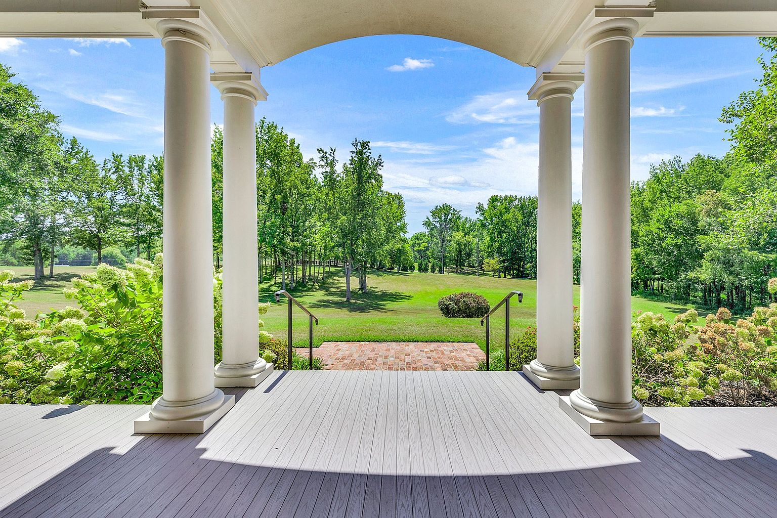 This image captures a serene view from a covered porch, framed by elegant white classical columns that lead the eye toward a lush, expansive backyard. The foreground features a clean, grey composite deck that transitions to a small brick landing with iron railings, overlooking a manicured lawn surrounded by mature trees. The perspective offers a peaceful, inviting atmosphere, highlighting the home's seamless connection to its natural surroundings.
