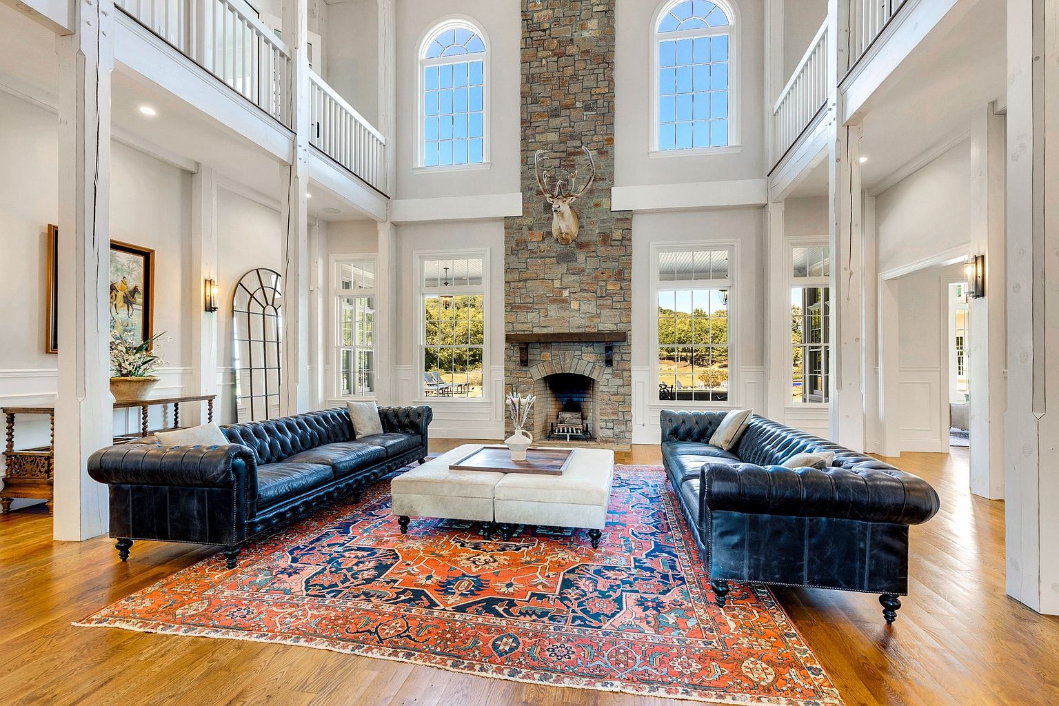 This is a grand living room featuring two large, tufted leather sofas facing each other across a patterned rug. A stone fireplace with a mounted deer head serves as a focal point, flanked by tall windows that provide ample natural light. The room's high ceilings and architectural details create a luxurious and spacious atmosphere.