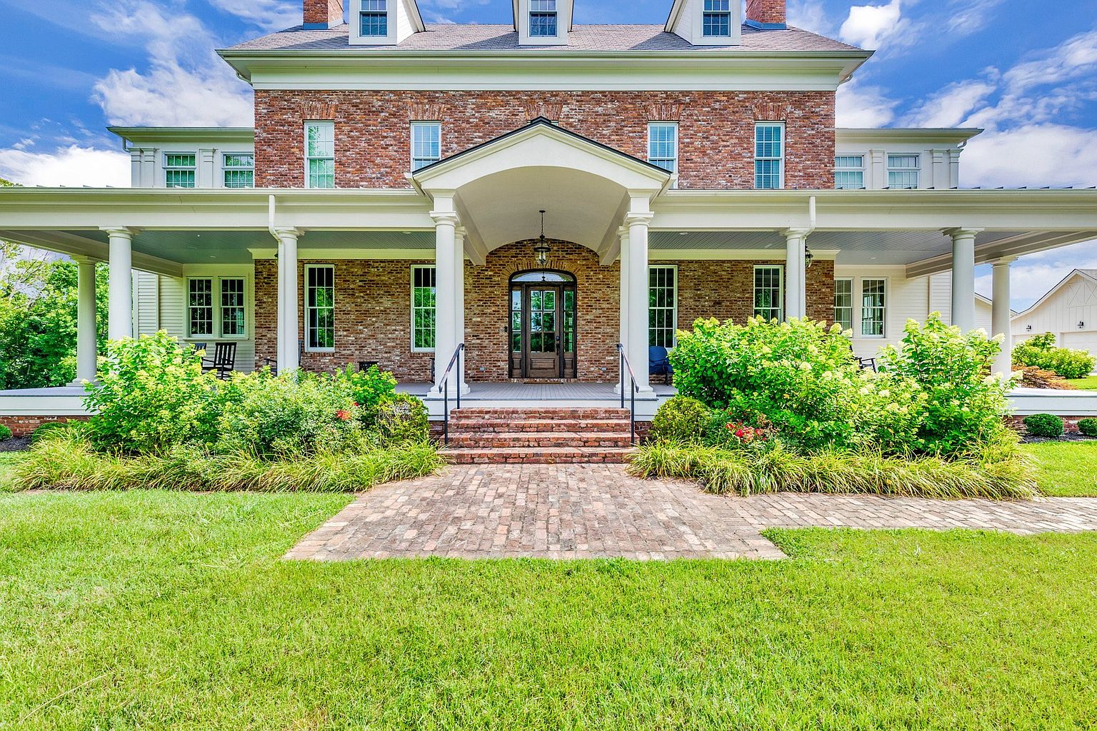 This stately brick home features a classic Southern-style wrap-around porch with white columns and a prominent, arched entryway. The symmetrical facade is accented by multiple dormer windows on the roof and lush, well-manicured landscaping that frames the brick walkway leading to the front door. The overall impression is one of timeless elegance, curb appeal, and traditional architectural charm.