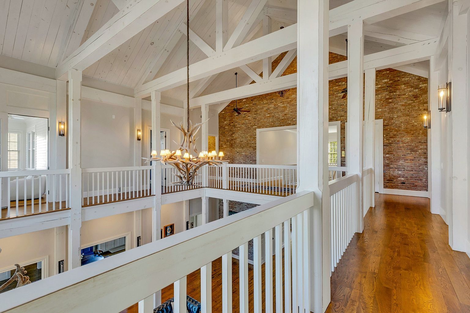 This interior shot showcases a spacious hallway with a multi-level design, featuring white wooden railings and support beams that create an open and airy feel. The hardwood flooring adds warmth, while the exposed brick wall provides a rustic contrast to the clean white elements. A unique antler chandelier hangs in the center, adding a touch of character to the space.