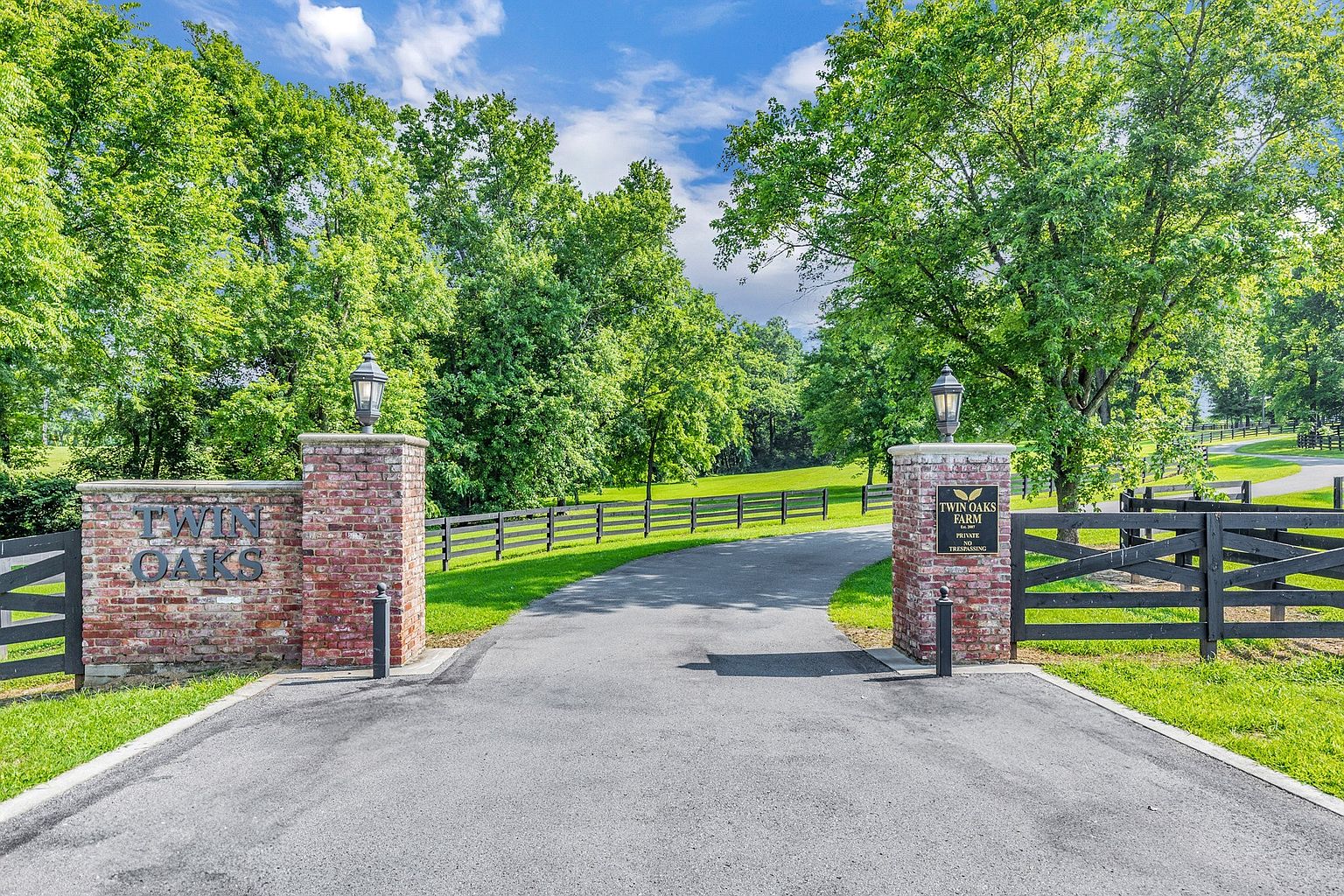 This image captures the grand, gated entrance to Twin Oaks Farm, featuring stately brick pillars topped with classic lanterns. A paved driveway leads through the open gate, framed by lush green trees and a traditional black wooden fence that stretches across the property. The scene conveys a sense of privacy, prestige, and well-maintained rural elegance.