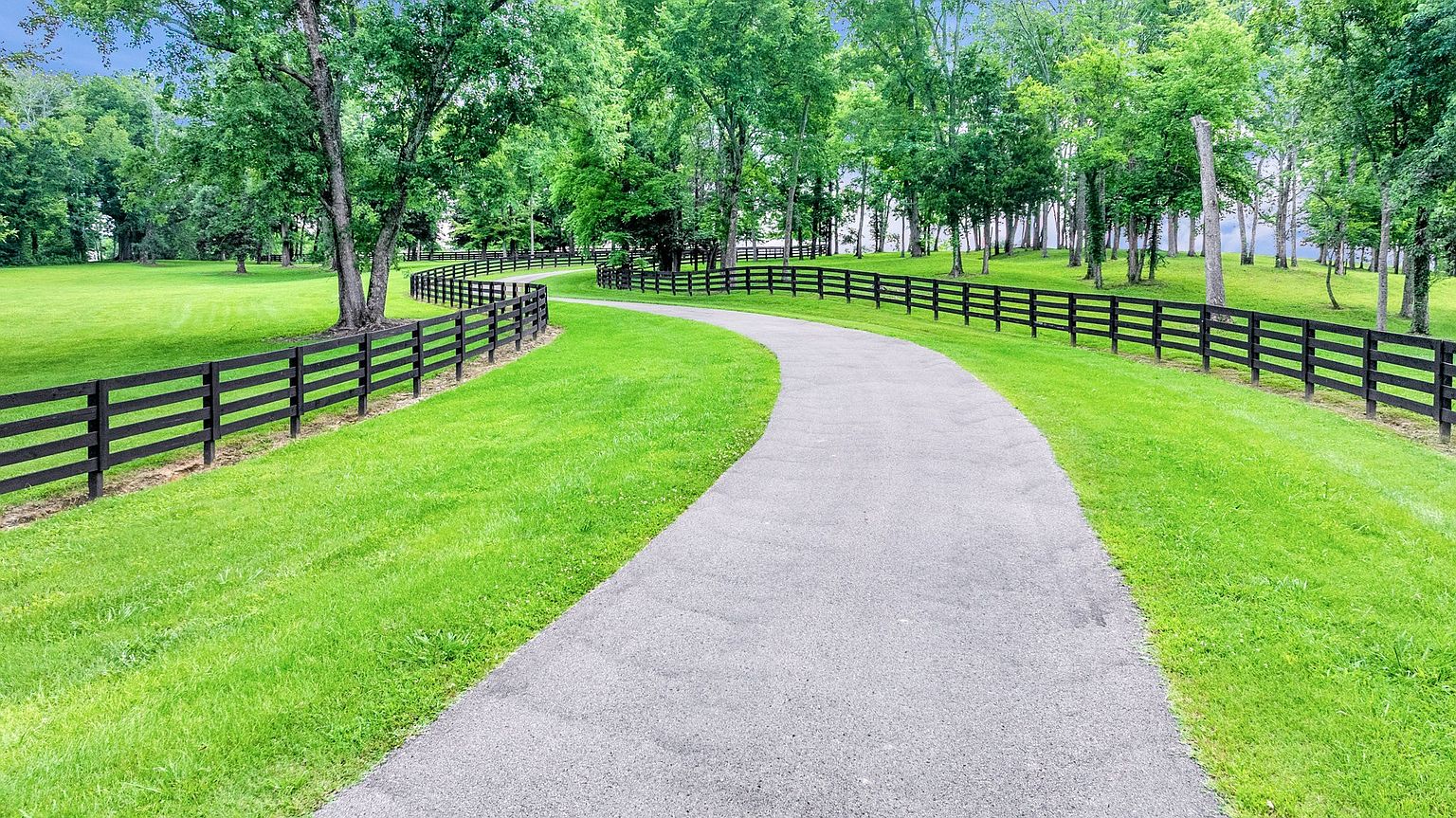 This scenic image captures a winding asphalt driveway leading through a lush, manicured green lawn framed by classic black wooden post-and-rail fencing. Mature, leafy trees line the background, creating a serene and expansive pastoral atmosphere. The perspective is a low-angle, eye-level shot that emphasizes the inviting curve of the path and the well-maintained landscape of the property.