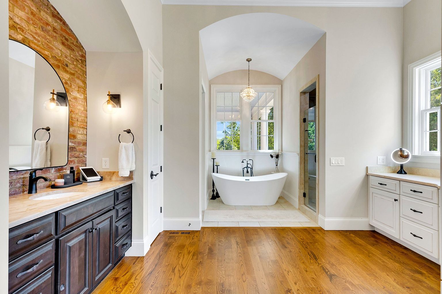 This is a luxurious primary bathroom featuring a freestanding bathtub positioned beneath an arched alcove with a window, allowing natural light to flood the space. The bathroom also includes a dark wood vanity with a stone countertop and a separate white vanity, complemented by hardwood flooring, creating a spa-like atmosphere. The perspective is from the doorway, showcasing the spaciousness and elegant design.