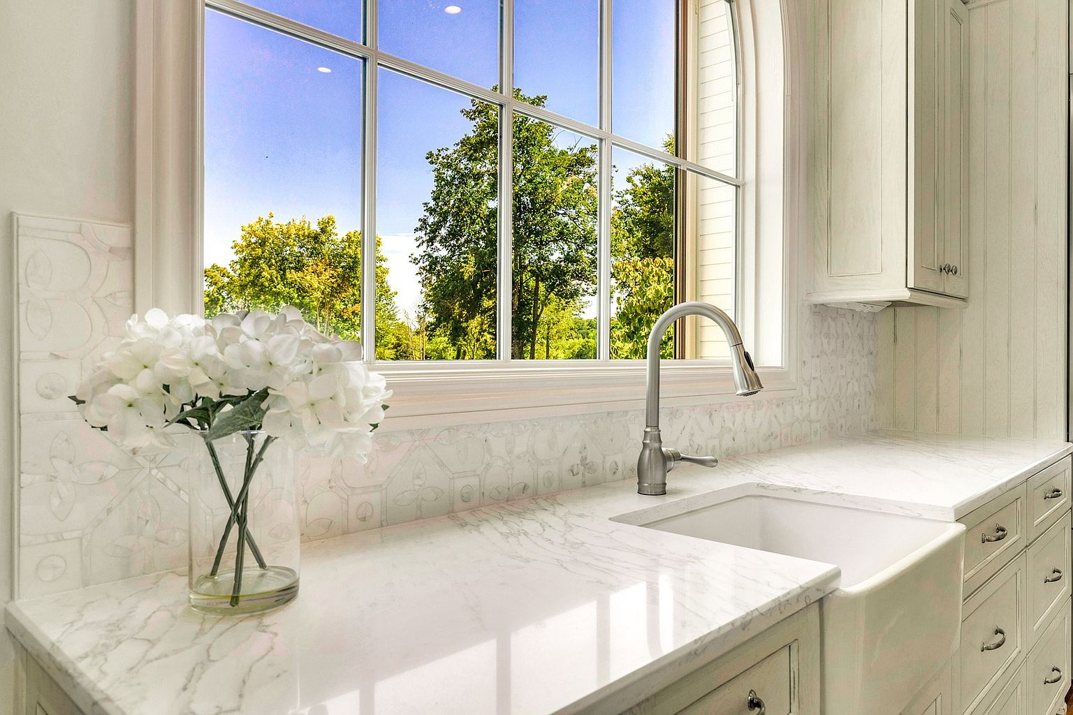 This elegant kitchen space features a classic farmhouse sink set into a pristine white marble countertop, complemented by intricate patterned tile backsplash. A large, arched window provides a bright, airy view of lush green trees, while white cabinetry with traditional hardware adds a timeless, sophisticated touch. The scene is accented by a clear glass vase filled with white hydrangeas, creating a serene and inviting atmosphere.