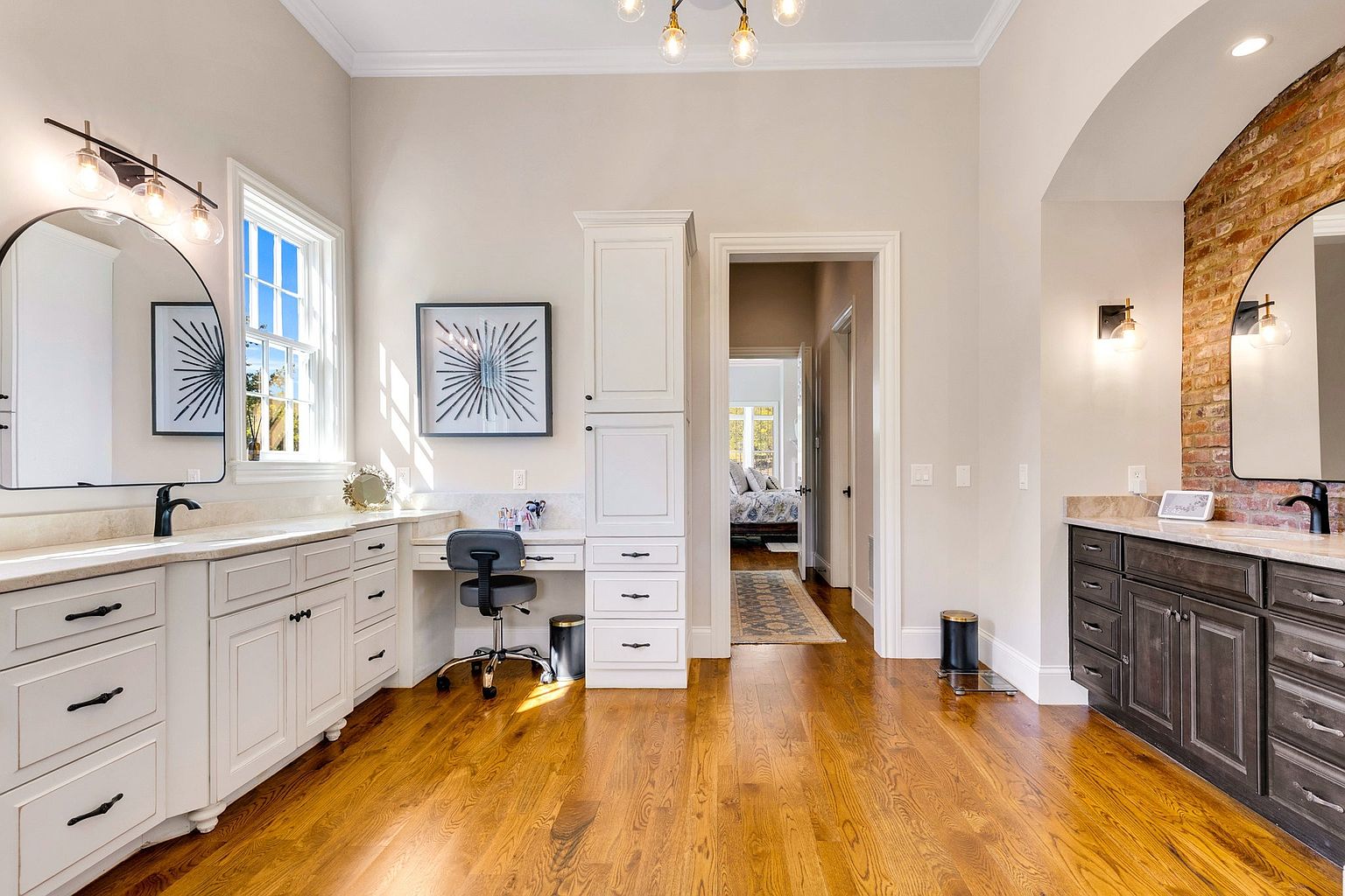 This spacious primary bathroom features a dual-vanity layout with contrasting white and dark wood cabinetry, topped with elegant stone countertops. The room includes a dedicated makeup vanity area, arched mirrors, and a warm, inviting atmosphere enhanced by hardwood flooring and a brick accent wall. A central doorway leads into an adjacent bedroom, creating a seamless flow between living spaces.