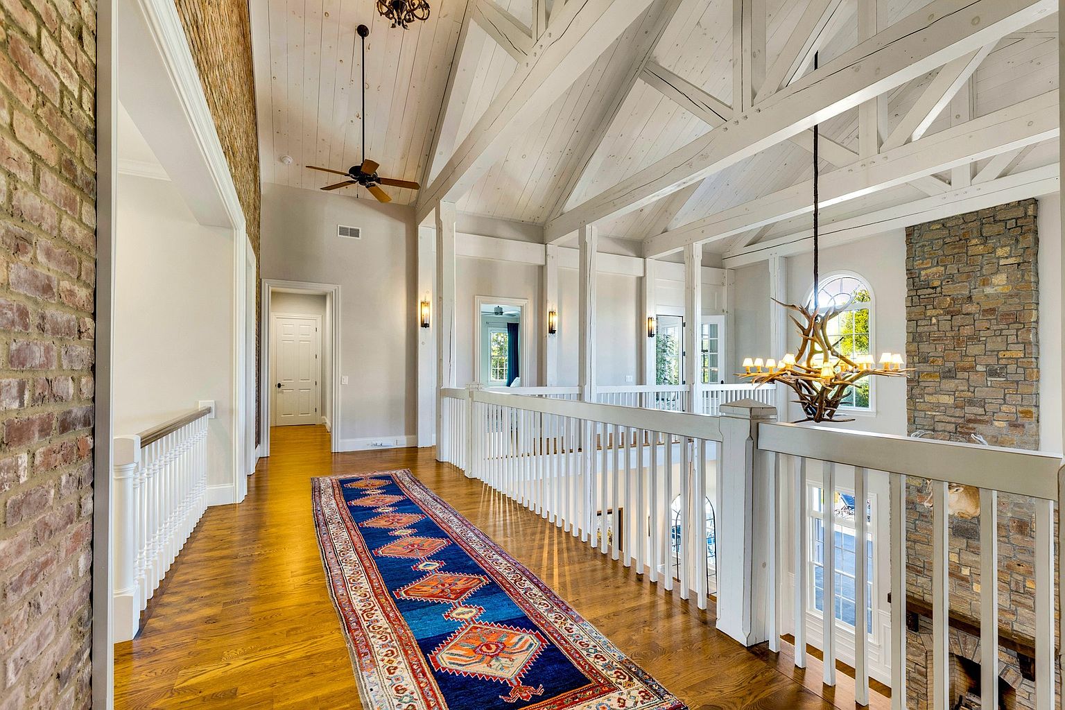 This elegant second-floor landing features a rustic-chic aesthetic with exposed white wooden trusses, a brick accent wall, and warm hardwood flooring. A vibrant, traditional-style runner rug adds a pop of color, while a unique antler chandelier serves as a striking focal point overlooking the space below. The open-railing design creates an airy, expansive feel that connects the upper level to the main living area.