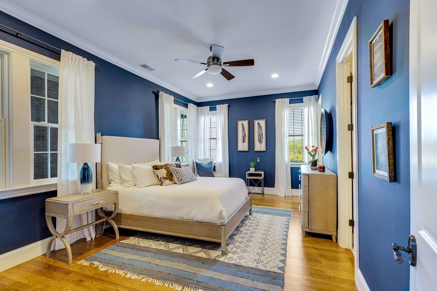 This elegant primary bedroom features deep navy blue walls that contrast beautifully with a light-toned upholstered bed frame and matching wooden nightstands. The room is bathed in natural light from multiple windows dressed in crisp white curtains, while a patterned area rug adds texture to the warm hardwood floors. A ceiling fan and tasteful wall art complete the sophisticated, serene atmosphere of this well-appointed space.