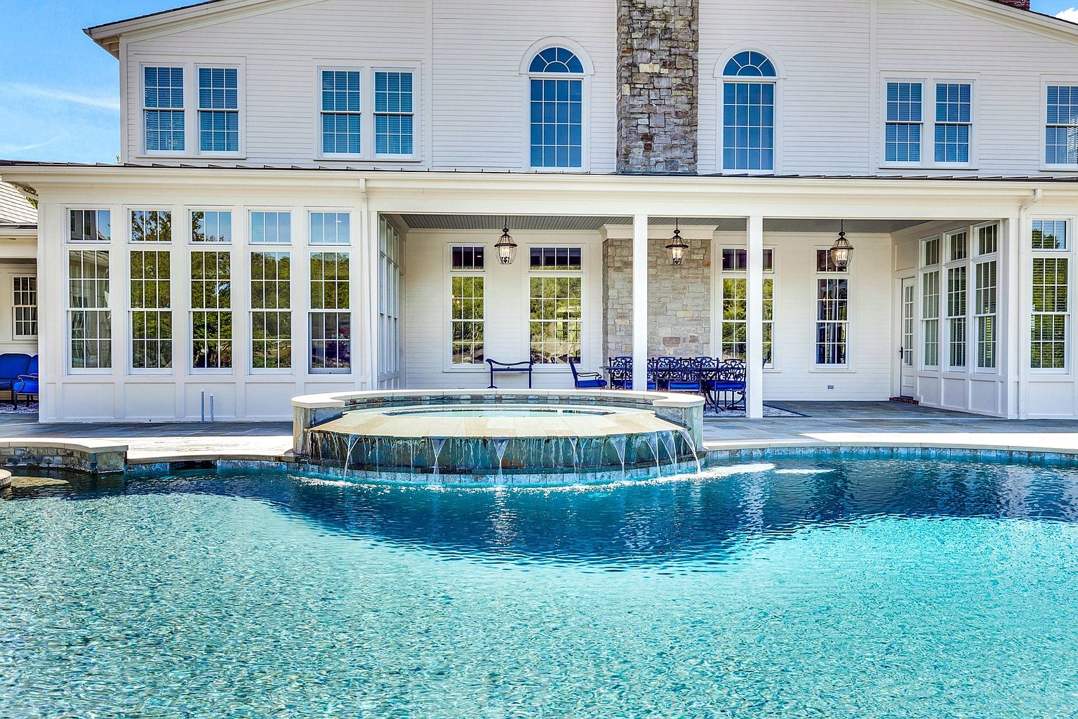 This stunning rear exterior view showcases a luxurious swimming pool with an integrated stone-clad spa that features a cascading waterfall edge. The white-sided home provides a classic, elegant backdrop, complete with a covered patio area furnished for outdoor dining and relaxation. The scene is bathed in bright, natural sunlight, emphasizing the pristine blue water and the sophisticated architectural design of the residence.