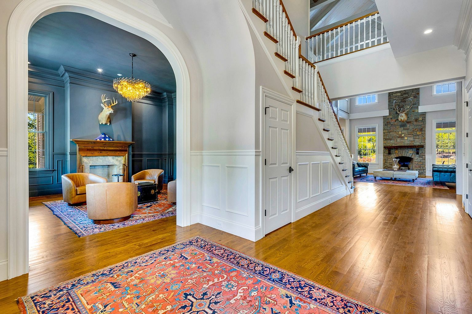 This grand foyer features a striking contrast between a moody, dark blue sitting room on the left and a bright, open-concept living area on the right. A classic staircase with white railings leads to an upper landing, while rich hardwood floors and intricate wainscoting add a sense of traditional elegance. The perspective captures the depth of the home, highlighting the architectural transition between the intimate, cozy den and the expansive, light-filled main living space.
