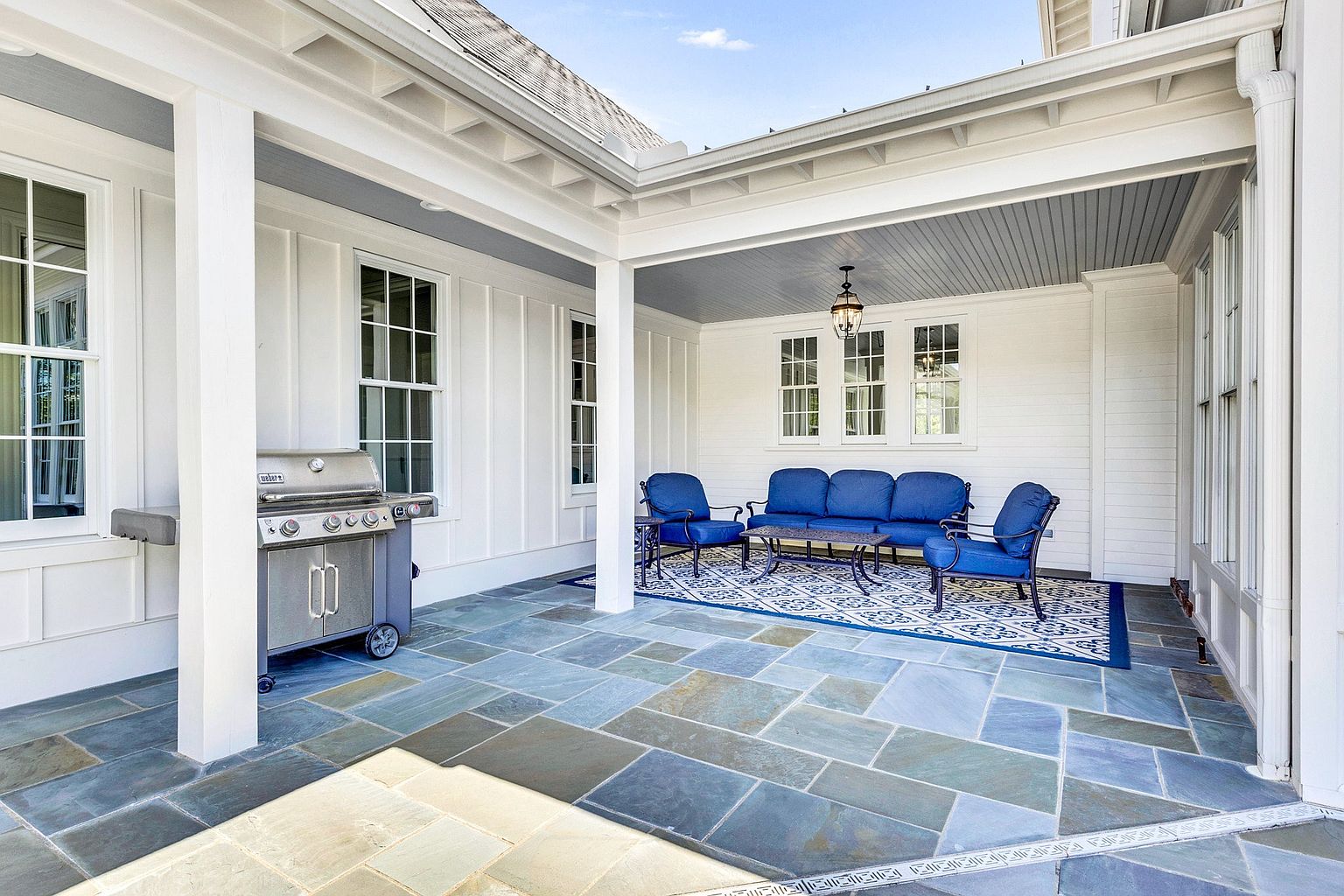 This inviting covered patio features elegant blue stone flooring and crisp white board-and-batten siding, creating a bright and airy outdoor living space. The area is furnished with a comfortable blue sofa set arranged on a patterned area rug, complemented by a stainless steel Weber grill. The perspective is a wide, eye-level shot that captures the seamless transition between the indoor-style comfort and the open-air architectural design.