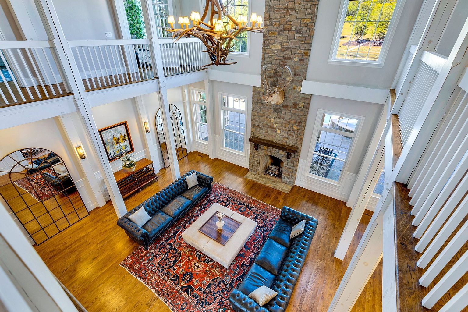 This high-angle, cinematic shot captures a grand, double-height living room characterized by a rustic-luxury aesthetic. The space features a massive stone fireplace adorned with a deer mount, a striking antler chandelier, and two dark leather tufted sofas arranged around a large, light-colored ottoman on a vibrant Persian-style rug. The room is framed by white wooden railings from the upper level, emphasizing the open, airy architecture and the warm, polished hardwood floors.