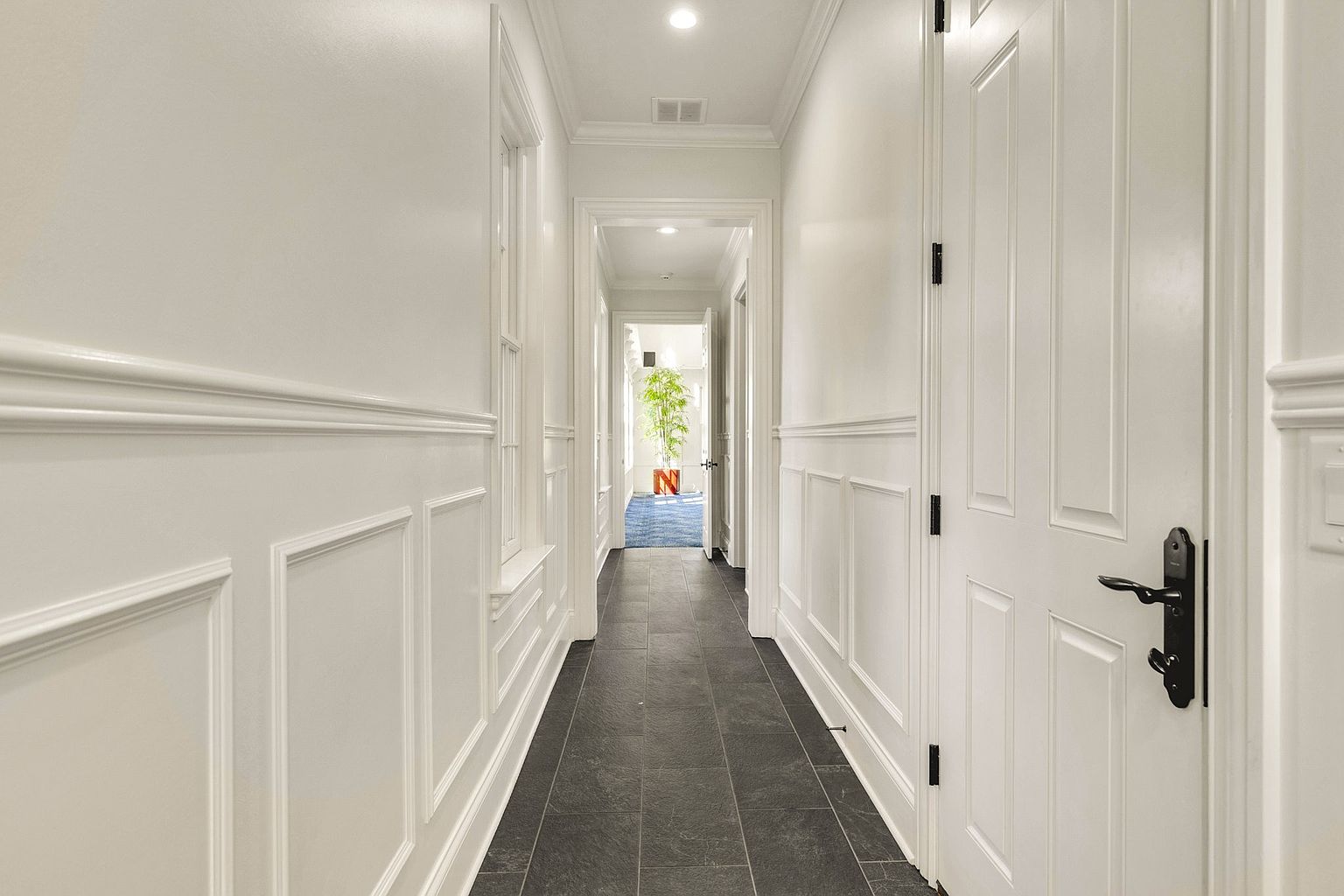 This elegant, long hallway features crisp white wainscoting and trim, creating a bright and sophisticated atmosphere. The dark, slate-toned tile flooring provides a striking contrast, while the perspective draws the eye toward a sunlit doorway at the end of the corridor. The clean lines and classic architectural details offer a polished, high-end aesthetic suitable for a luxury residence.